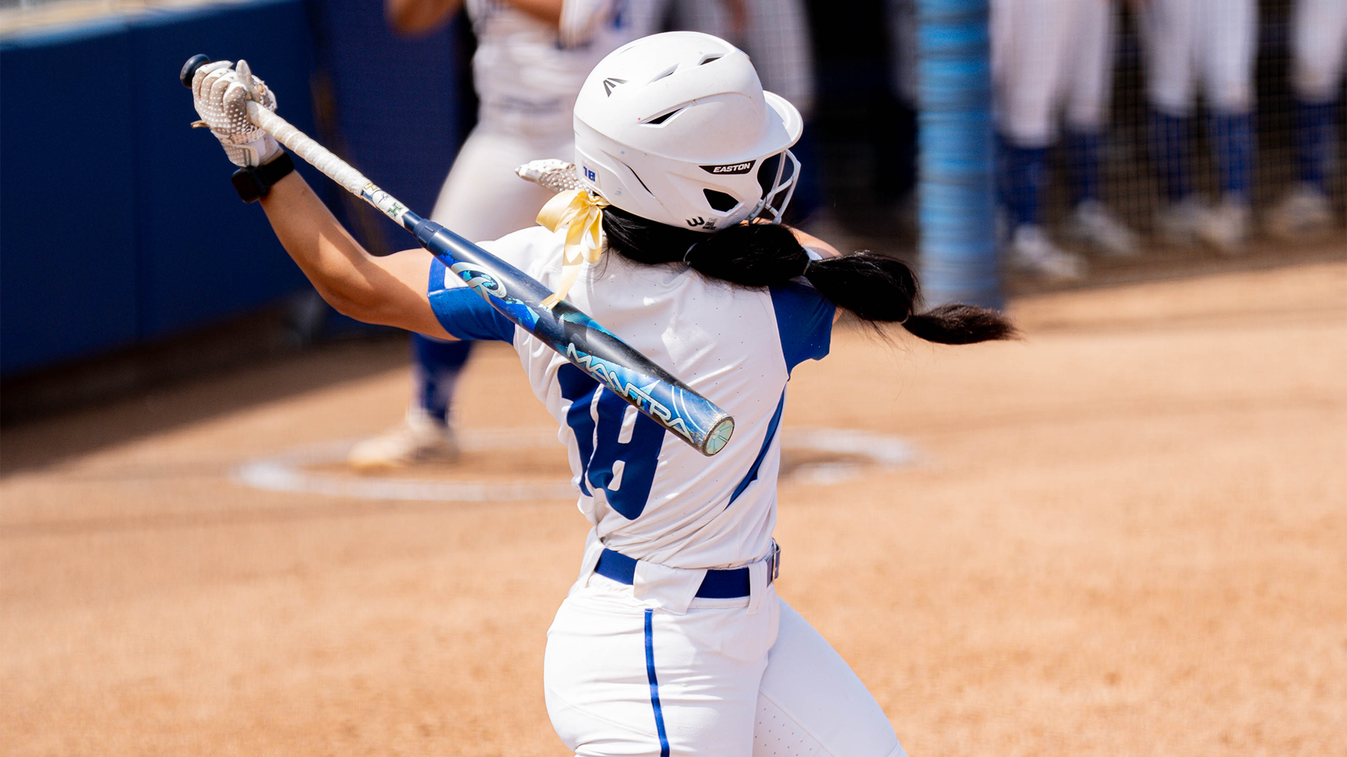 Bella Fuentes (#18) finishes her swing before stepping into the batters box. Her back is turned to the camera with her number showing and the bat lined across her back. 
