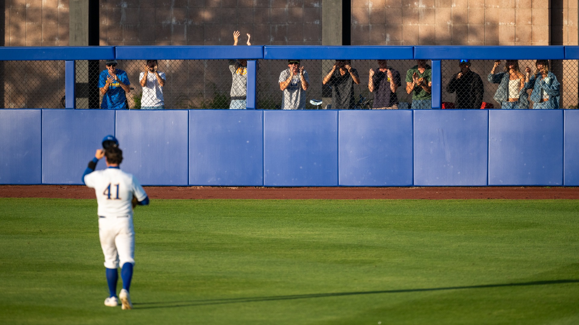 A group of Gaucho fans peer through chain-link fence at the top of the right field wall. In the foreground, #41 Noah Karliner faces the fans and tips his cap in acknowledgement 