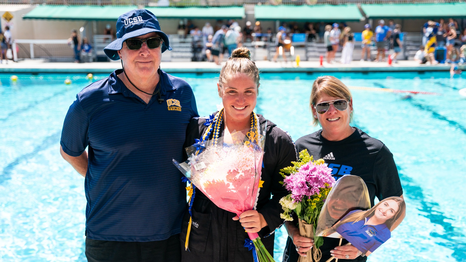 Cami Mras (#18) stands between her parents, Jim (left) and Kim (right) during her Senior Day ceremony. Cami and Kim both have bouquets of flowers in their hands. Kim is also holding cardboard cutouts of photos of Cami