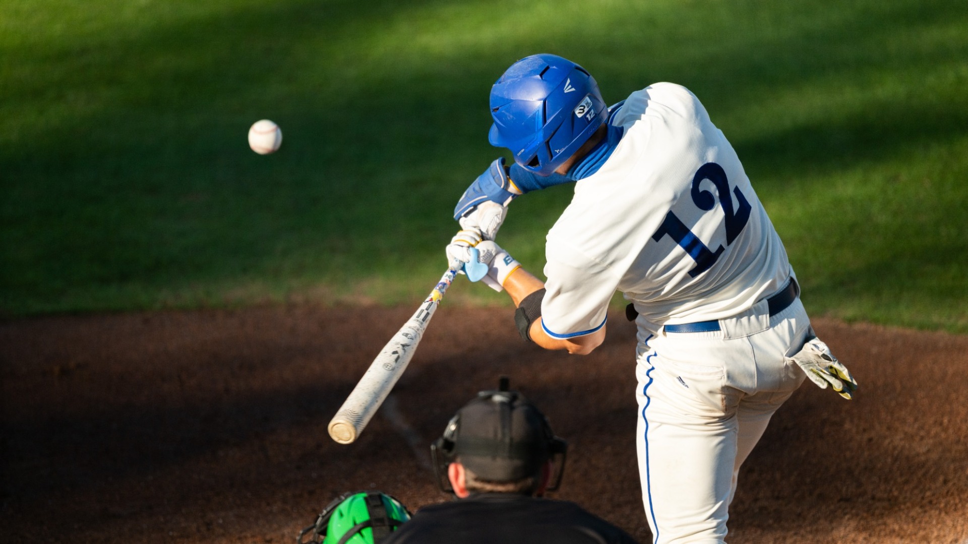 A photo of Nate Vargas (#12) batting, taken from behind as he swings at a pitch. Vargas' back is fully facing the camera as he swings through, and we can see the ball just in front of his bat.
