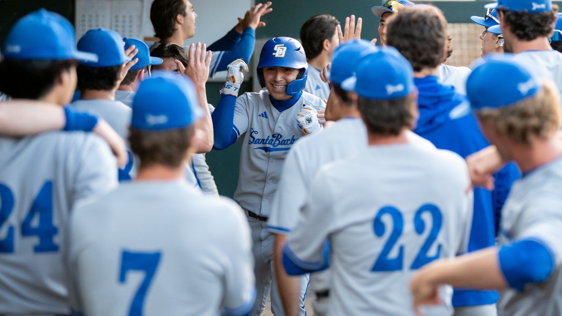#5 Jonathan Mendez grins under his batting helmet as teammates surround him with high-fives in the dugout after scoring a run