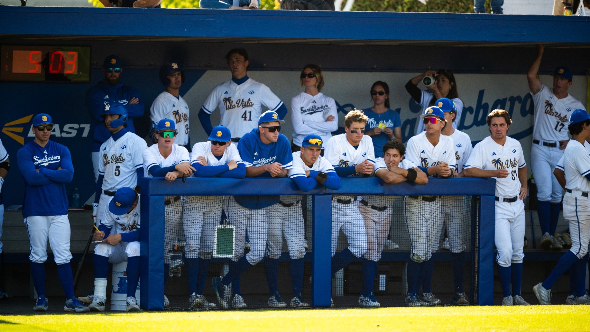 A number of Gaucho baseball players lean up against the railing of their dugout in the evening light. More teammates and staff stand on the bench behind them, making for two tiers of Gauchos in the dugout.