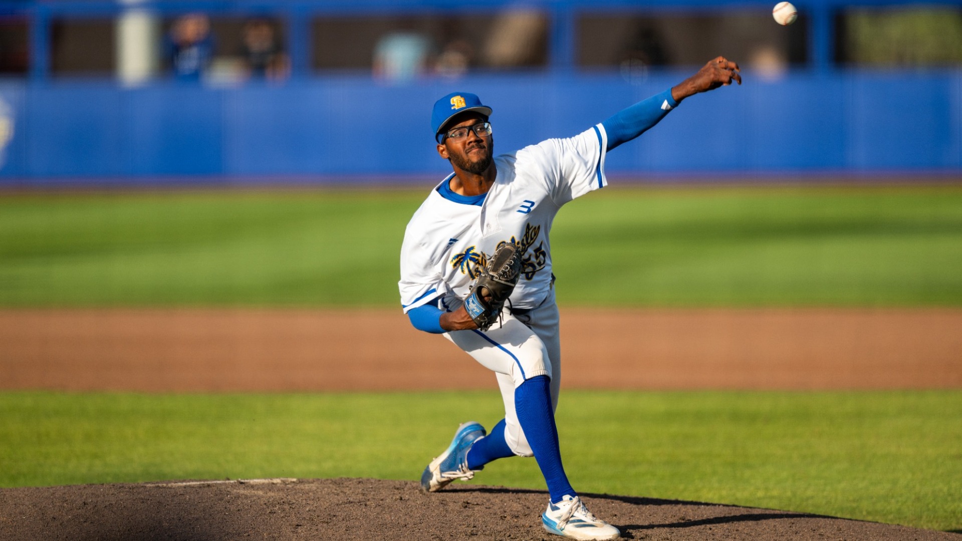 #55 Donovann Jackson delivers a pitch from the mound at Caesar Uyesaka Stadium. The ball has flown out of his hand and is in the top right corner of the frame, as we view Donovann from the third-base side of the field