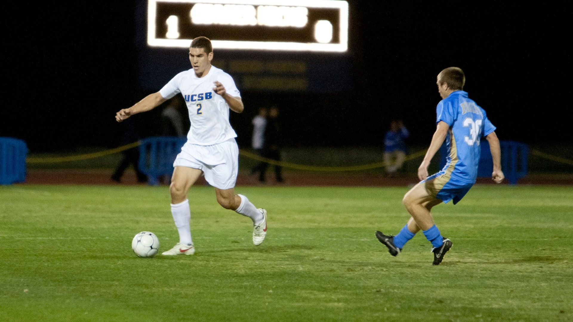 A photo of Michael Boxall playing for the Gauchos in 2009. He has the ball at his feet, facing toward the left side of the frame and preparing to dribble. A UCLA player is approaching him from the right side of the frame