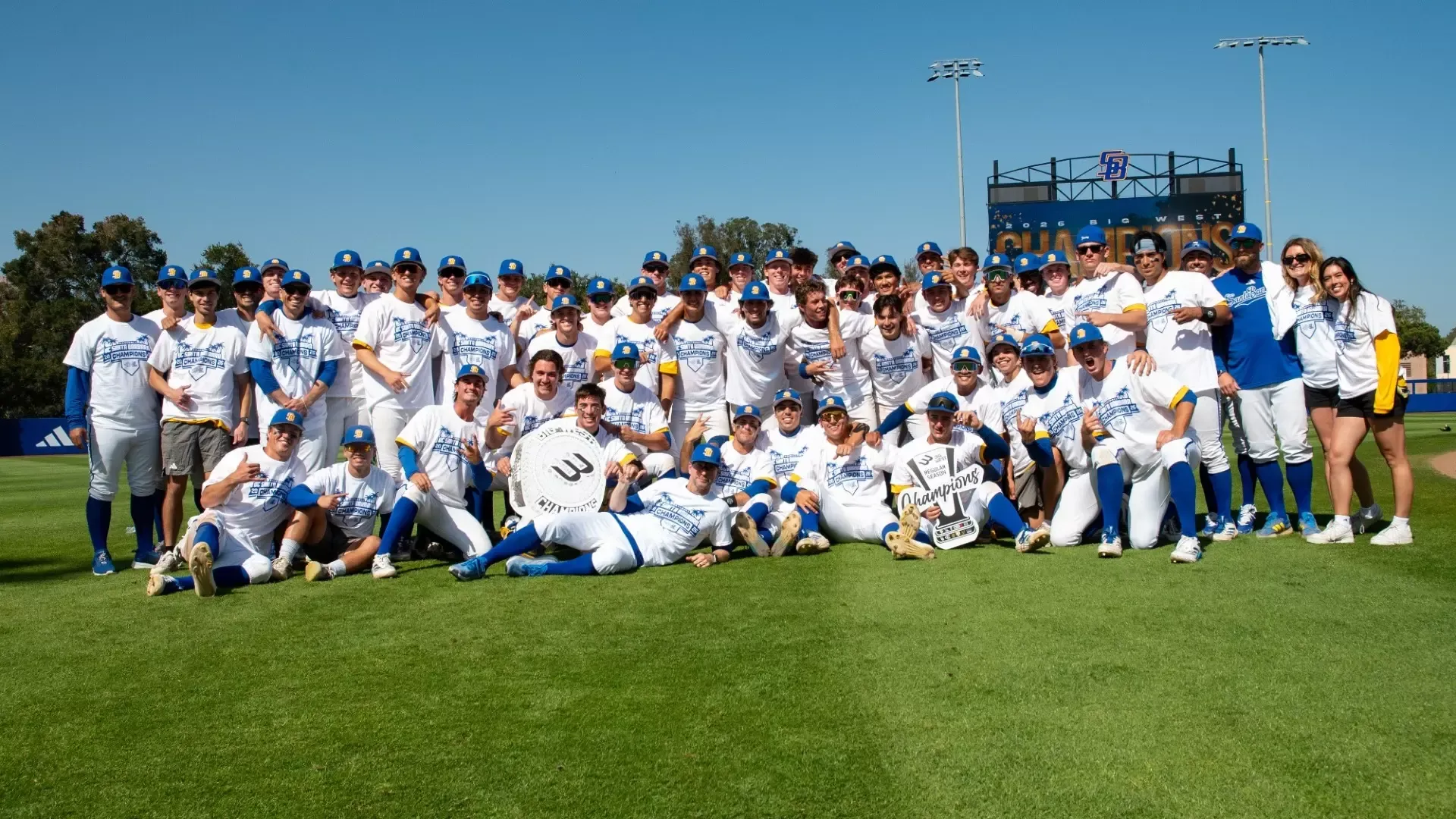 The UC Santa Barbara Baseball team, all wearing matching white Big West Regular Season Champions t-shirts, pose for a photo on the outfield grass at Caesar Uyesaka Stadium.