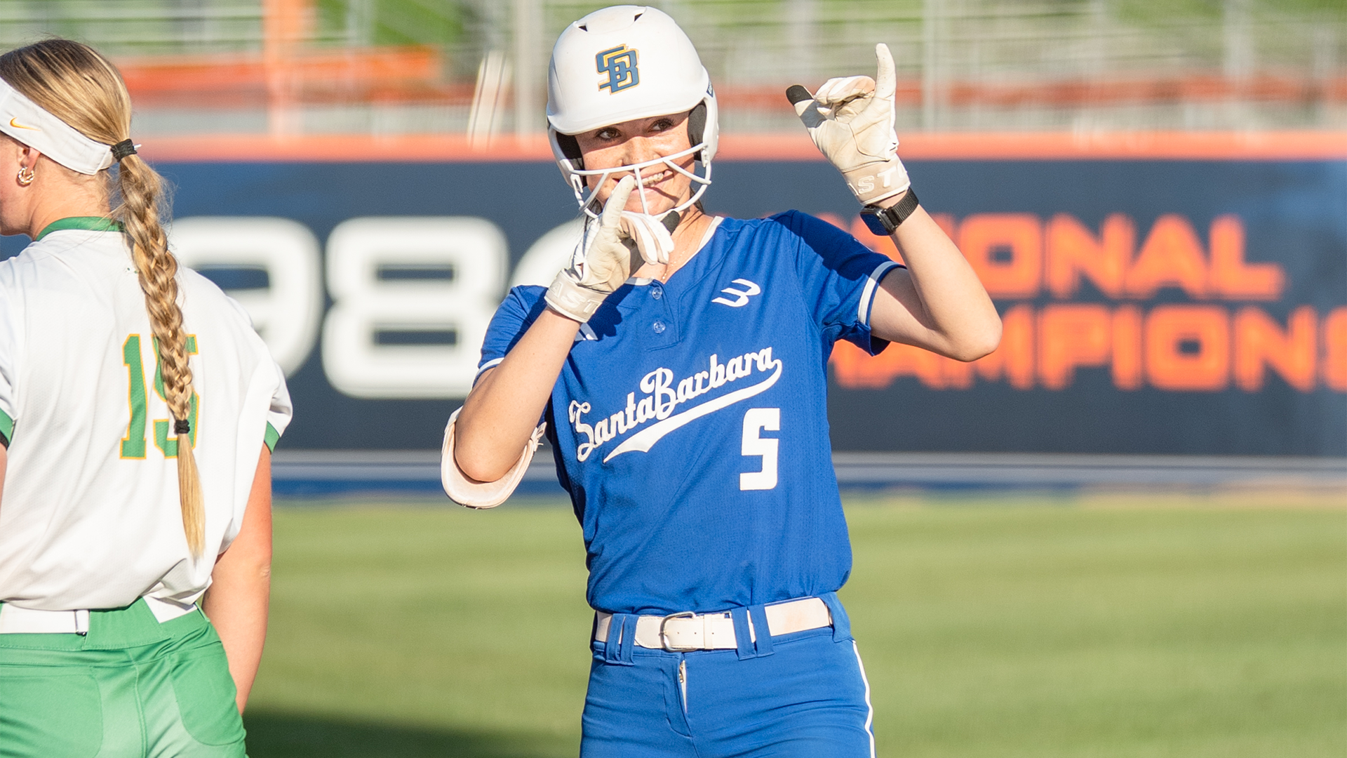 Dani Rauscher stands on first base after a hit. She is smiling and hold up hand gestures to the dugout.