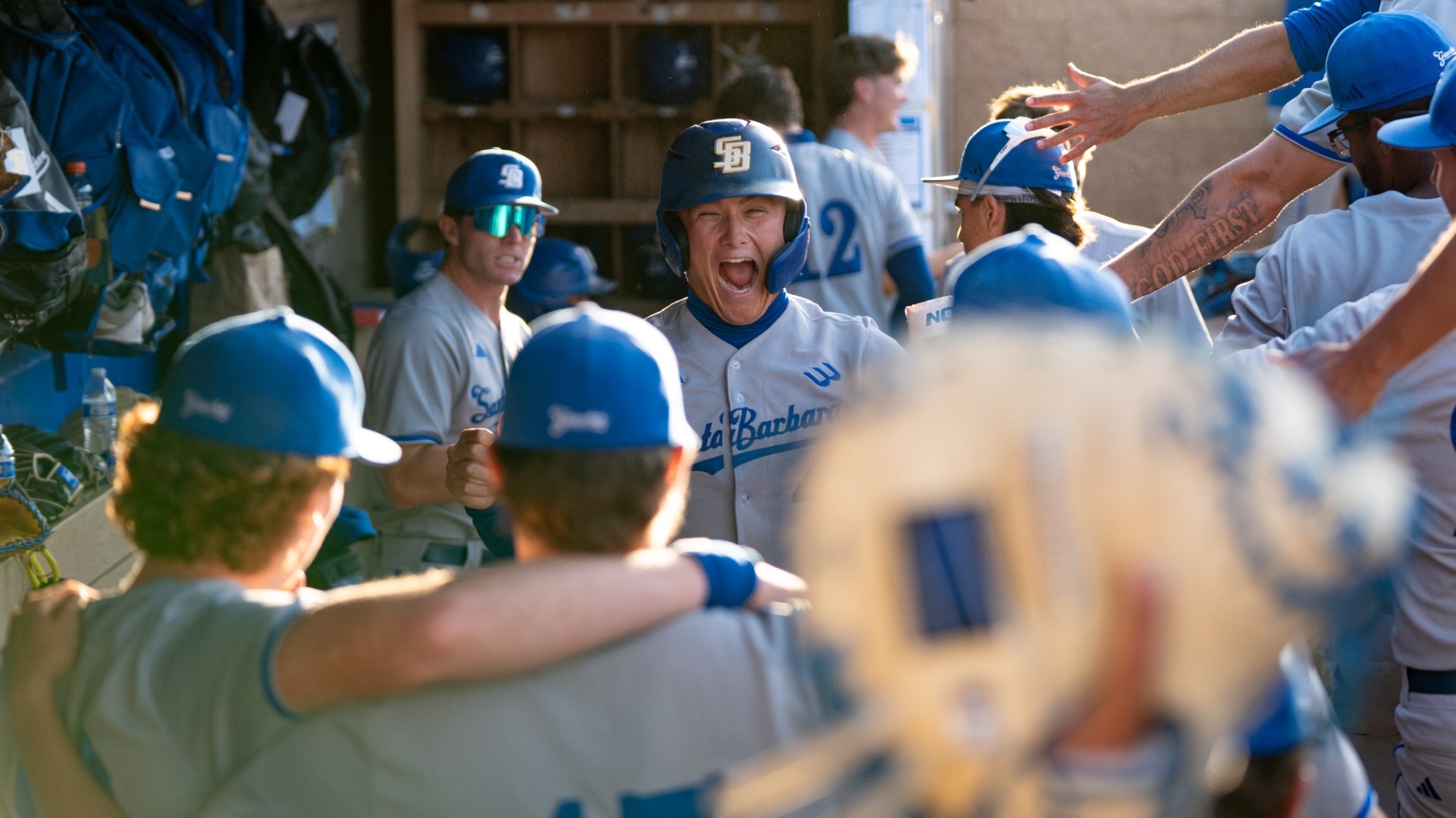 #41 Noah Karliner screams in celebration with his teammates in the dugout at Cal State Bakersfield. Out of focus in the foreground, we see two Gauchos waiting at the end of a line of high-fives to celebrate with their teammate