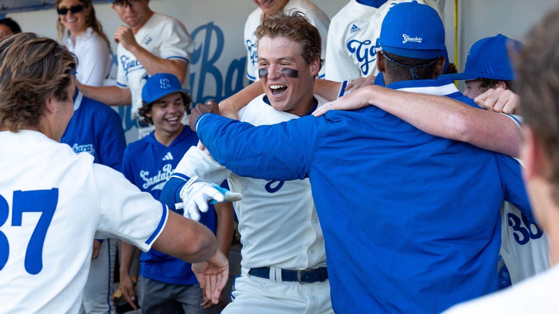 #26 William Vasseur celebrates with a teammate, wearing a blue jacket, in a half-hug, half-dance in the Gauchos' dugout. He flashes a toothy grin and looks towards a teammate half in frame