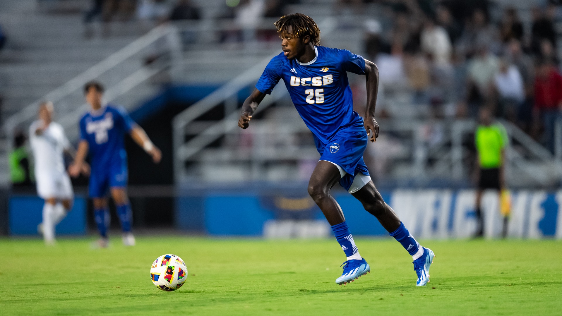 #26 Manu Duah dribbles the ball up field. He looks towards the left of the frame, eyeing his next pass as the ball sits in front of him, leaning forward to start running.