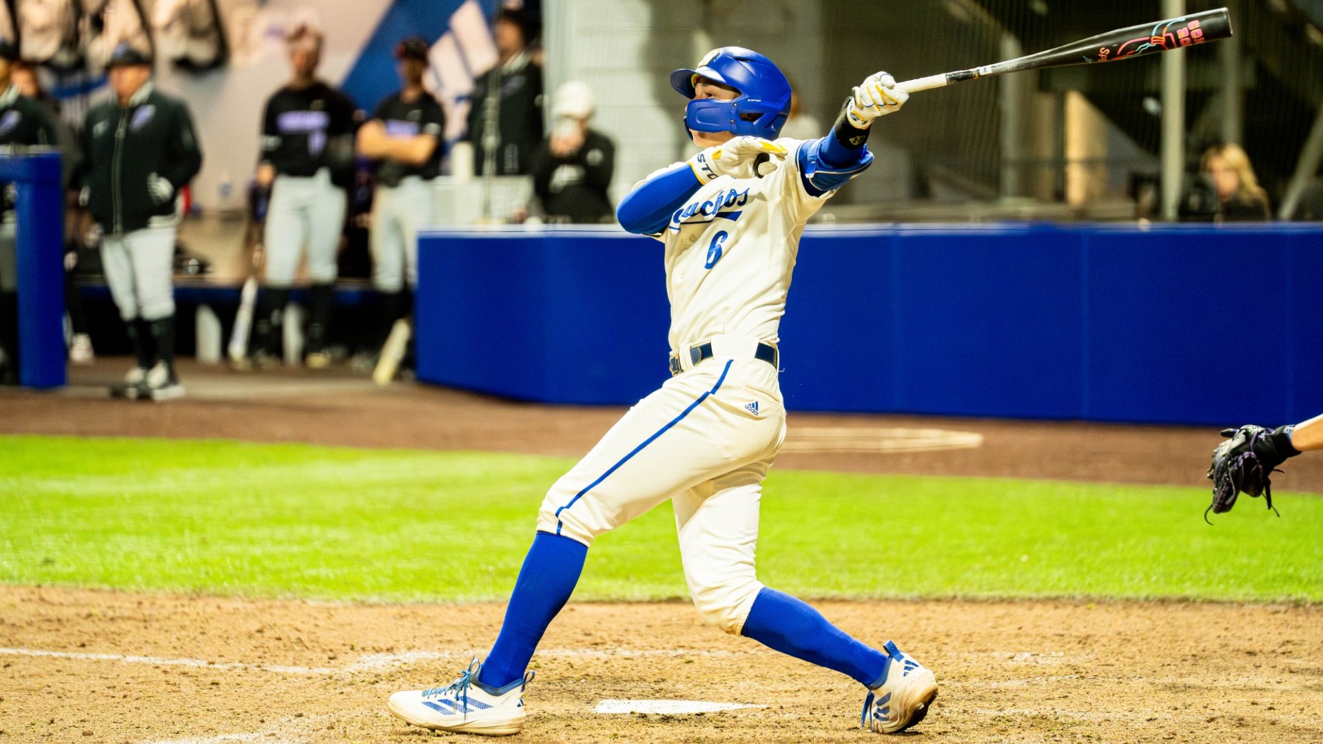 #6 Nick Husovsky follows through a big swing under the lights at Caesar Uyesaka Stadium. His right hand is low, off his bat as his body twists around, the bat extended high behind him.