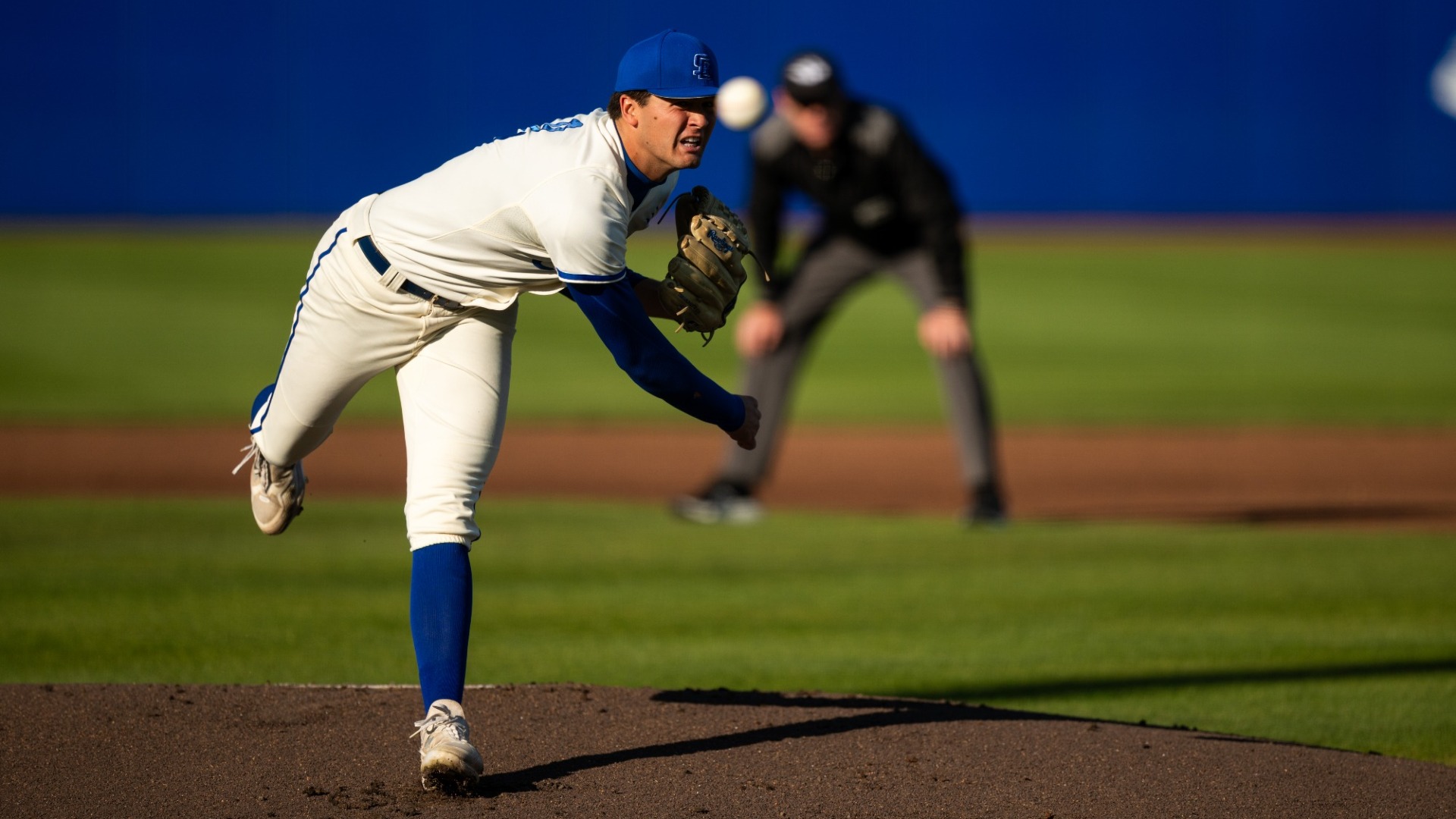 A view from behind home plate as Jackson Flora delivers a pitch at Caesar Uyesaka Stadium. He is bent over at the hip and we can see the ball just in front of him, fired out of his hand