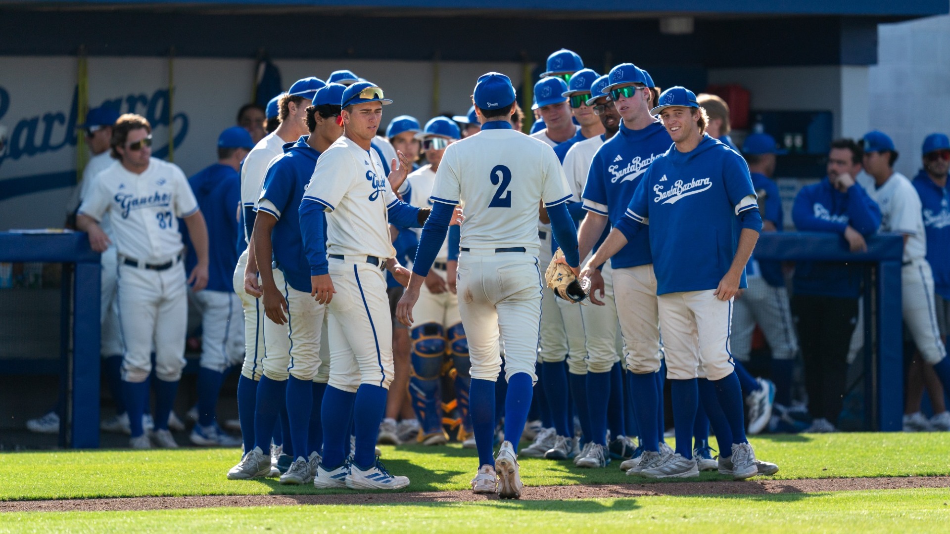 A view from across the field as Jackson Flora walks back into the UC Santa Barbara dugout. His teammates line up on both sides of him, smiling, to congratulate him on his work