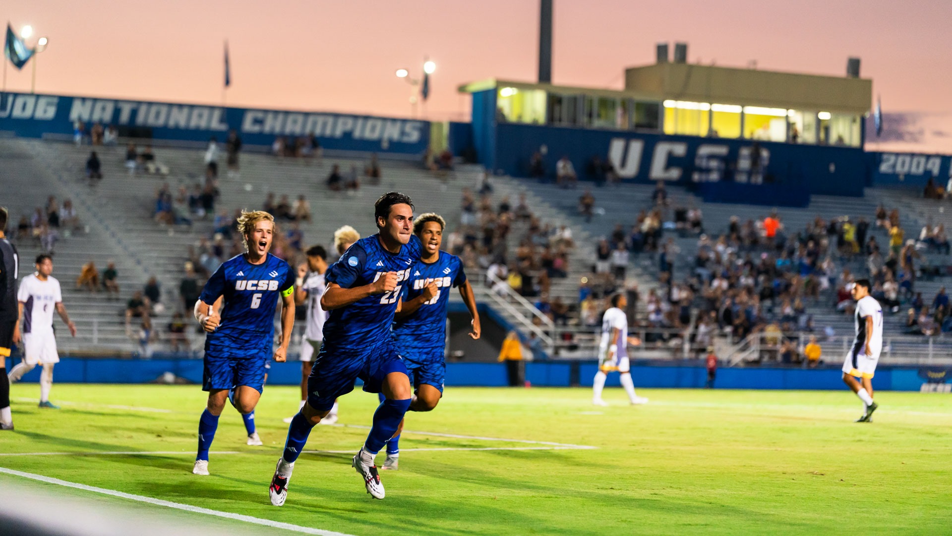 #23 Simone Ferrieri runs away from the goal in celebration after scoring. #6 Kalle Rahbek and #18 Tobi Gerber run behind him. Above the stands in the background, the sky is pink from the sunset