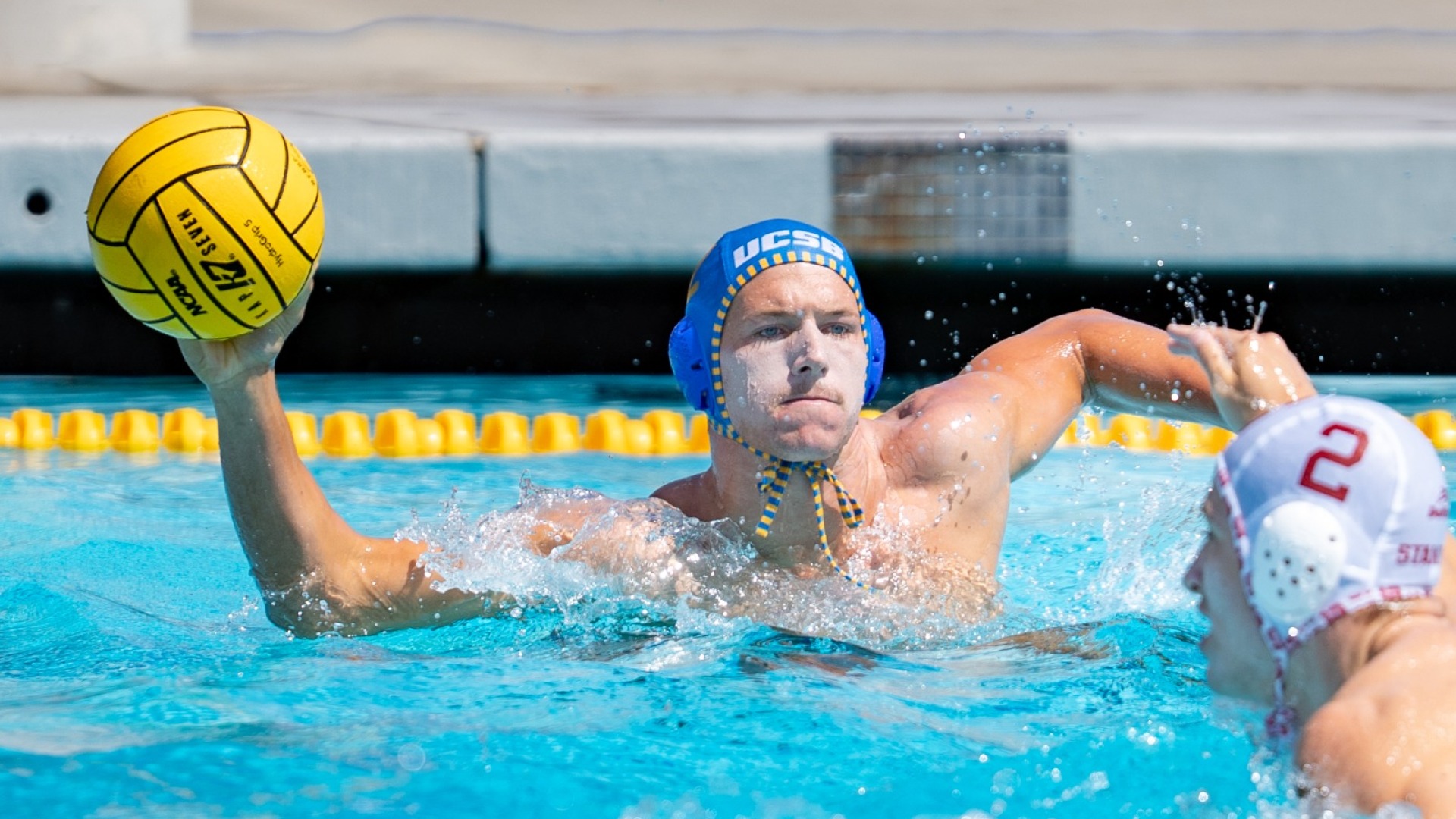 #12 Tyler Roller holds the ball in his right hand as he swims down the wide area of the pool, looking in towards the camera to find a teammate to pass to