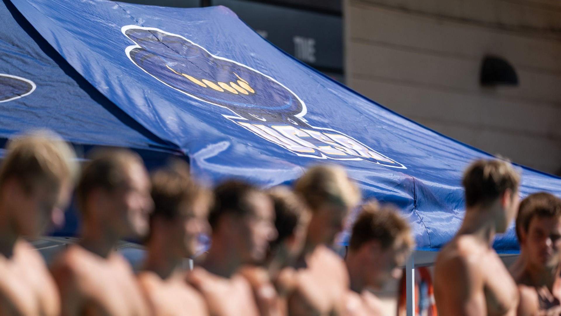 A blue UCSB-branded tent stands behind a line of water polo players on the pool deck at Santa Barbara High School