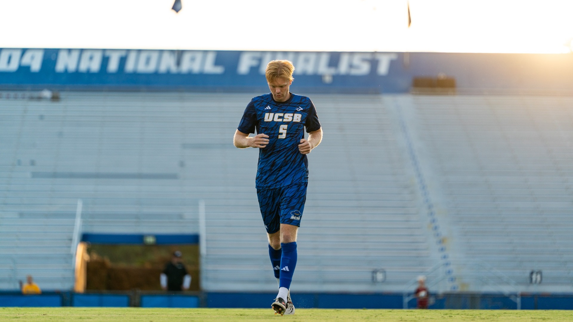 #5 Viggo Gustavsson jogs towards the camera, the sky a white gold in the evening light behind him. The 2004 National Finalist sign on the Harder Stadium wall is visible but out of focus behind him.