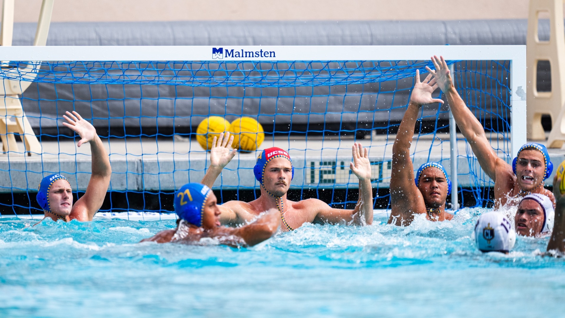 #1A Haakon Lacy readies himself in the goal, with his arms up at right angles out of the water. On either side of him, Gaucho teammates raise an arm to try and block an impending shot