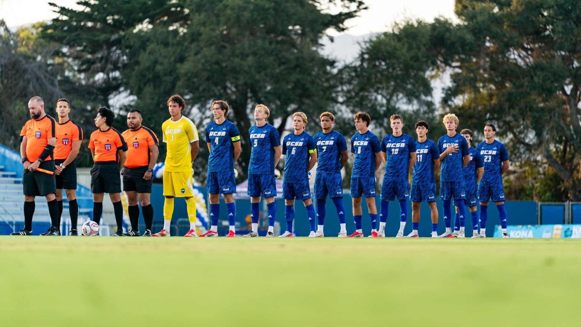 The UC Santa Barbara Men's Soccer team's starting 11 stands in a line at midfield during the playing of the national anthem.