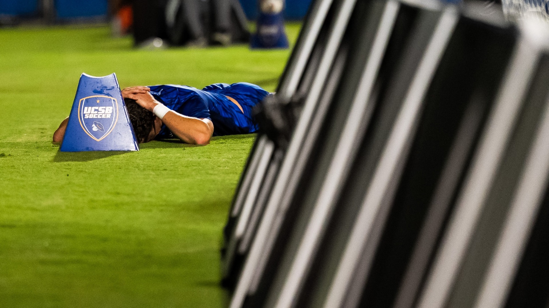 A Gaucho Men's Soccer player lays on the ground behind one of the goals and advertising boards at Harder Stadium. His head is partially hidden behind a pitchside ball holder, which has the UCSB Soccer shield logo on it.