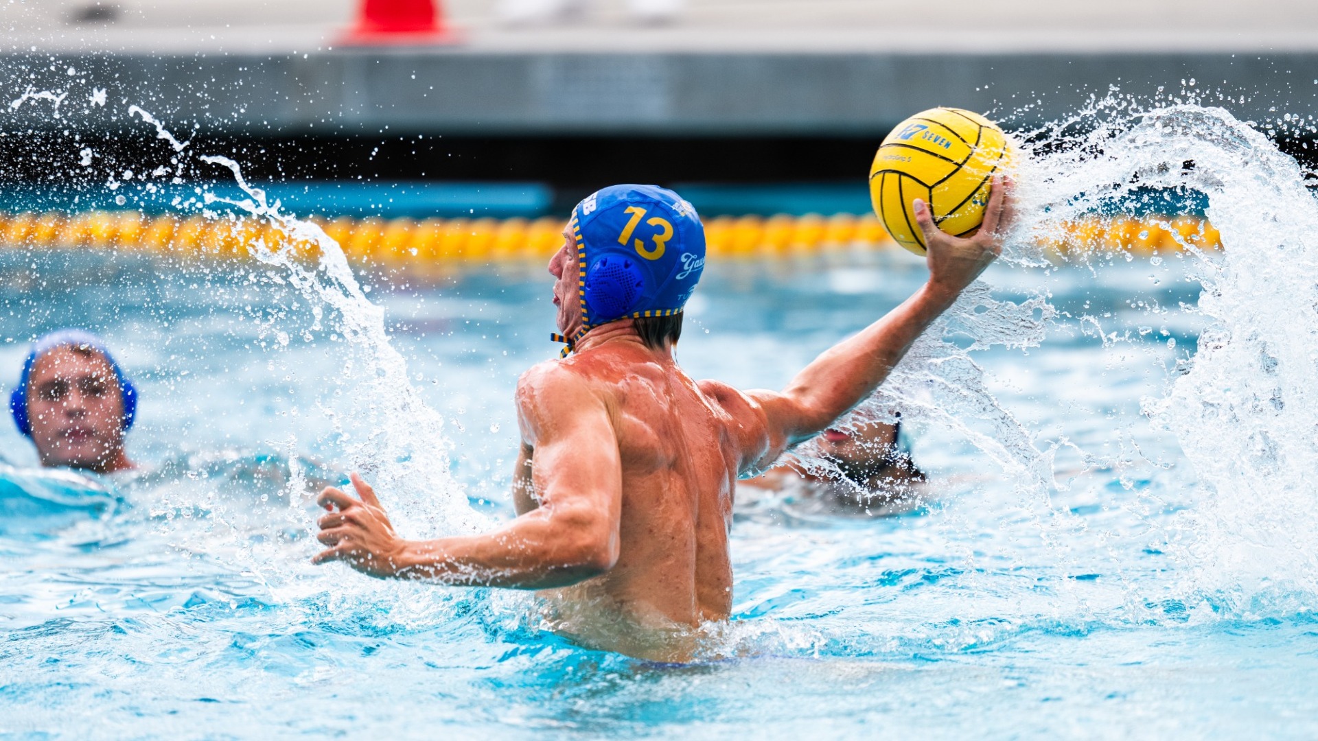 A profile view as #13 Kyle Franks raises his entire upper body out of the water and cocks his right arm back to shoot the ball. A rooster tail of water flows off the ball to the right of the frame as Kyle lifts it