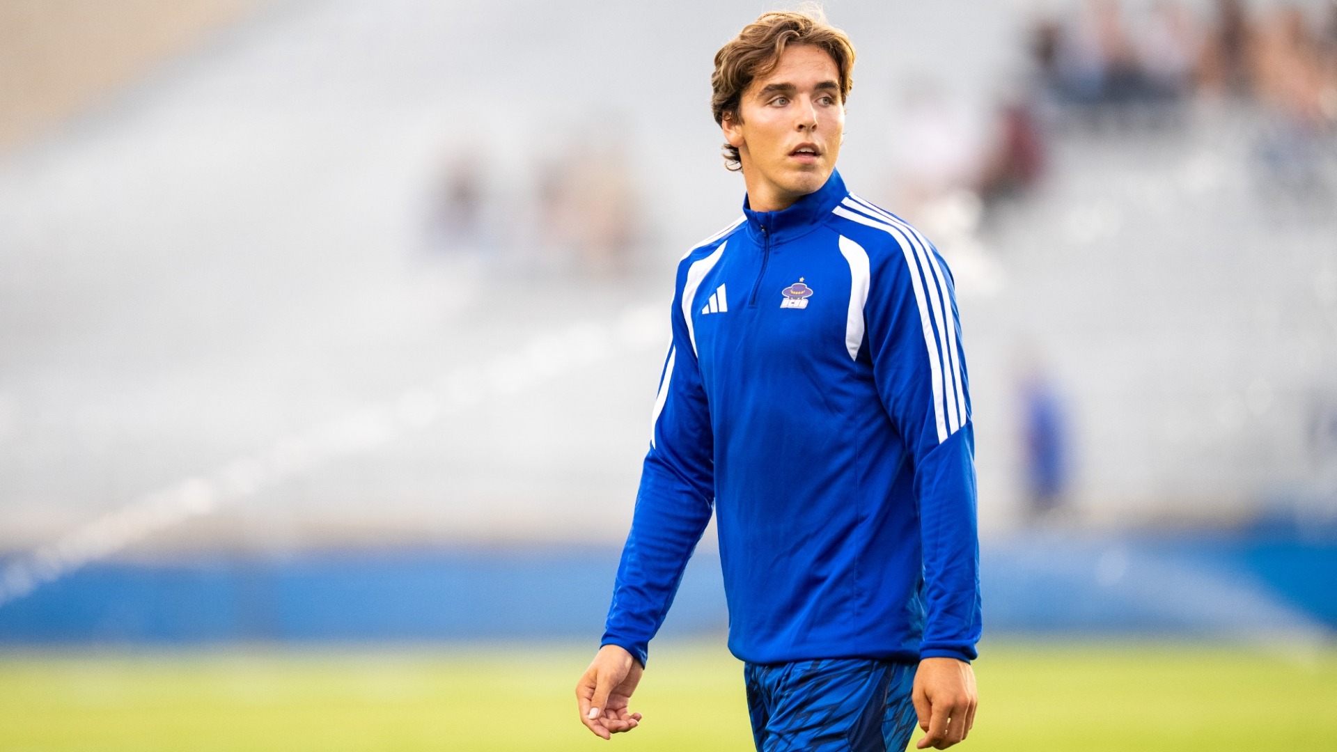 Gunnar Franke strides across the field in his blue warm-up jacket and match shorts, bathed in golden evening sunlight