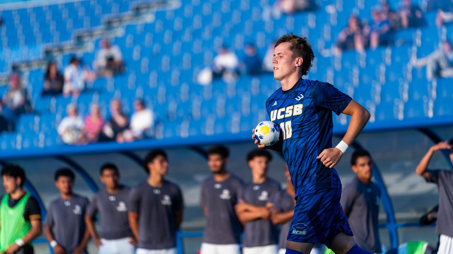 #10 Steinar Bjornsson runs onto the field at Harder Stadium in the evening sun. The blue seats in the stands are bathed in the evening glow behind him