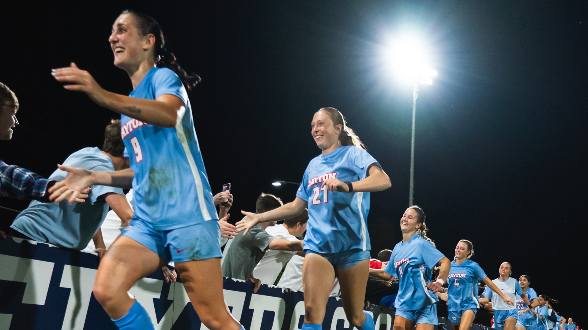 University of Dayton women’s soccer players wearing light blue uniforms smile and high-five fans along the sideline after a game under the lights. The players, including numbers 9 and 21 in the foreground, extend their arms toward an enthusiastic crowd leaning over the barrier. The scene is lit by a bright stadium light against the dark night sky, capturing a celebratory postgame moment.