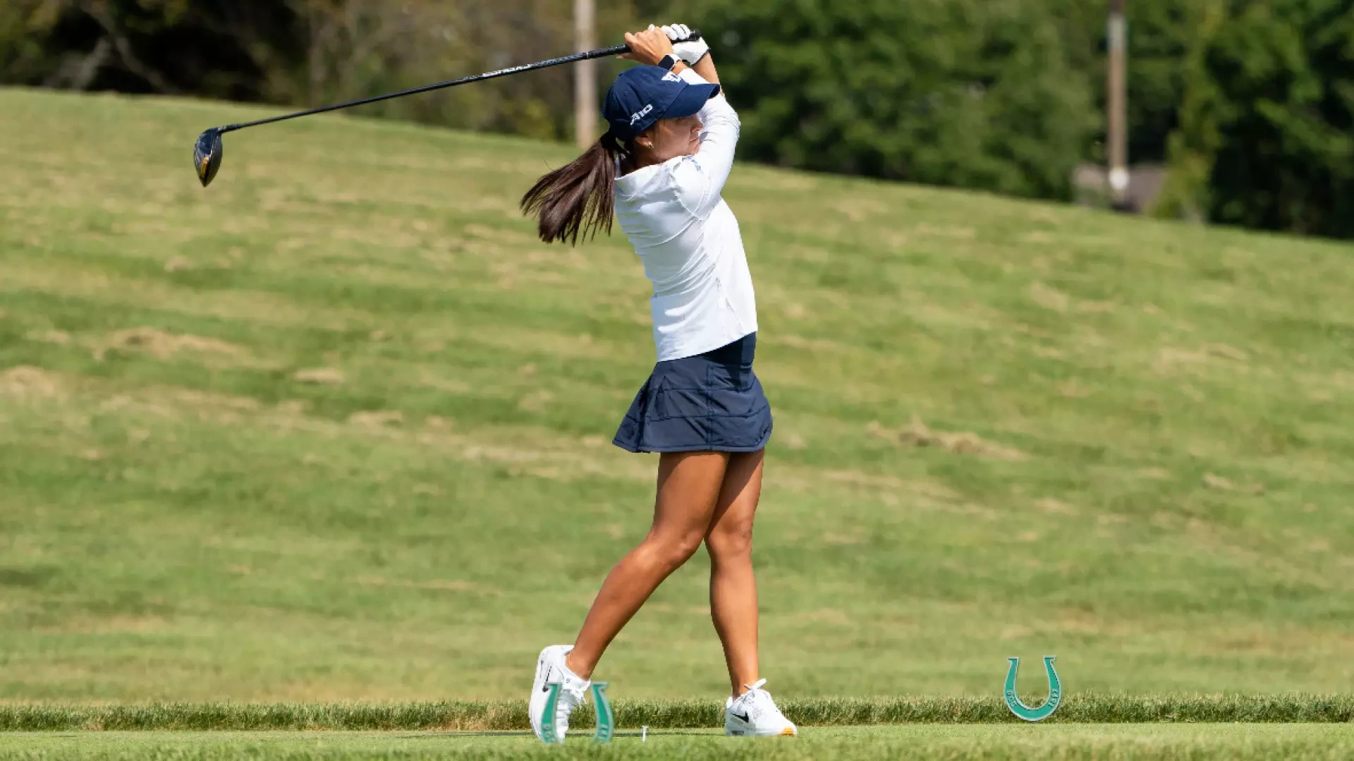 Women's golfer Christina Pfefferkorn, wearing a white top, blue hat and blue skirt, in her backswing in a tee box at Dayton Country Club during a practice round