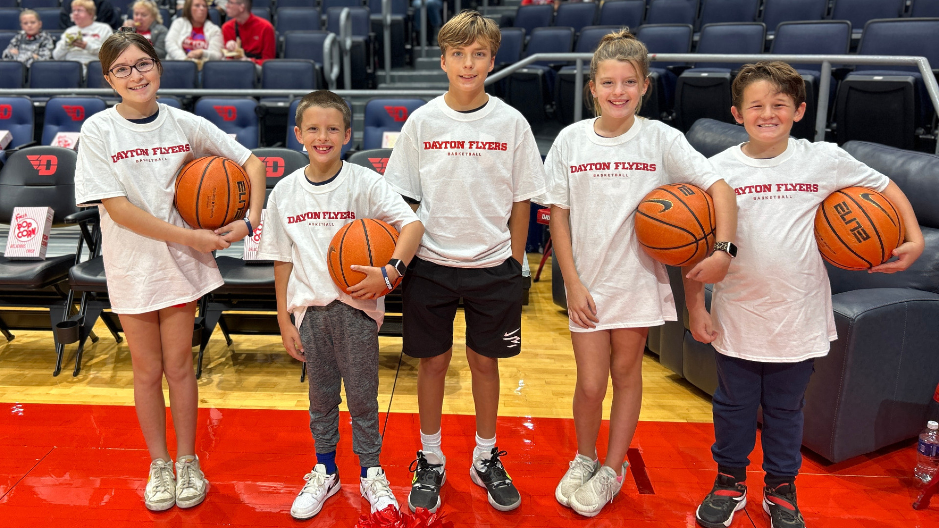Several kids wearing a white Ball Kids t-shirt stand on the sideline of a men's basketball game at UD Arena holding basketballs