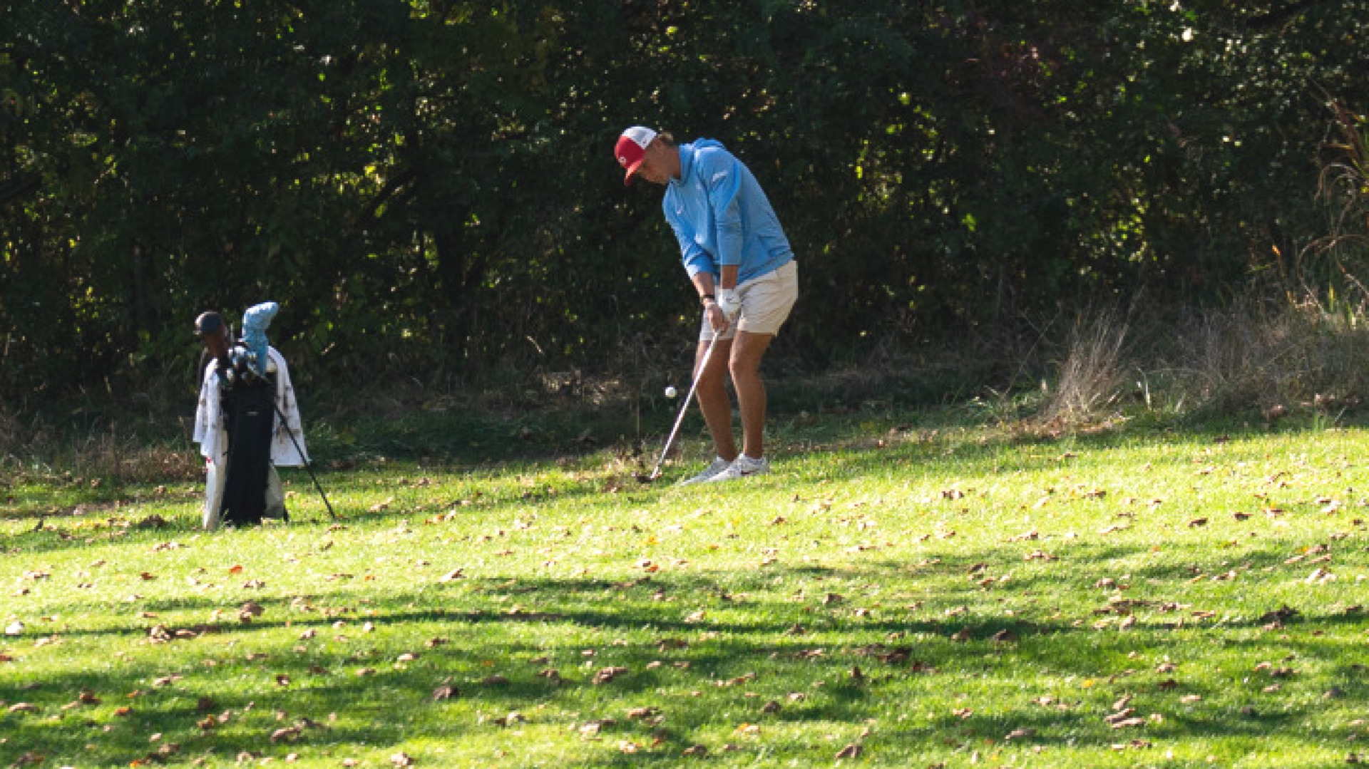 Men's golfer T.J. Kreusch, wearing a red and white hit, chapel blue pullover and white shorts, hitting out of the rough at NCR Country Club