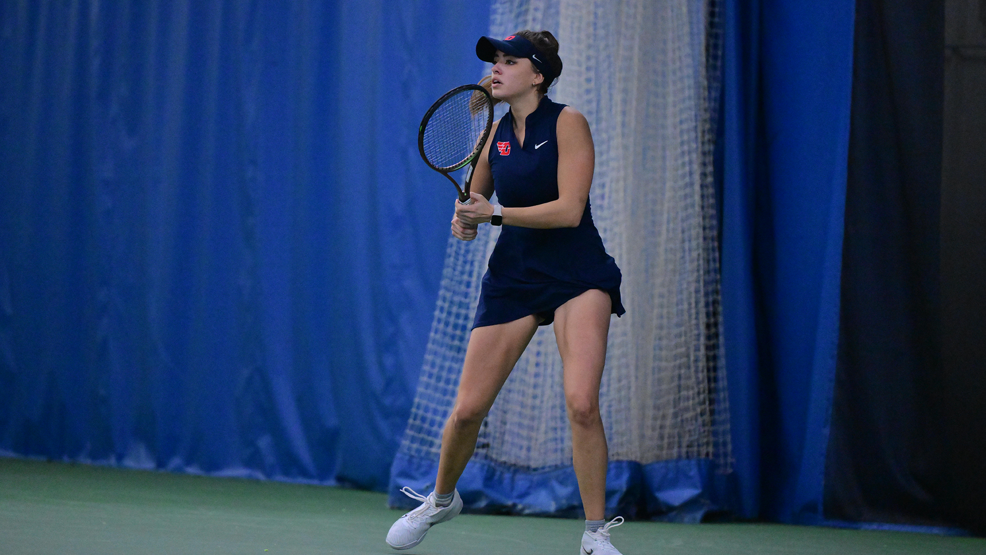Montserrat Sierra stands ready with a tennis racquet in her right hand. She is in a navy uniform and wearing a navy visor.