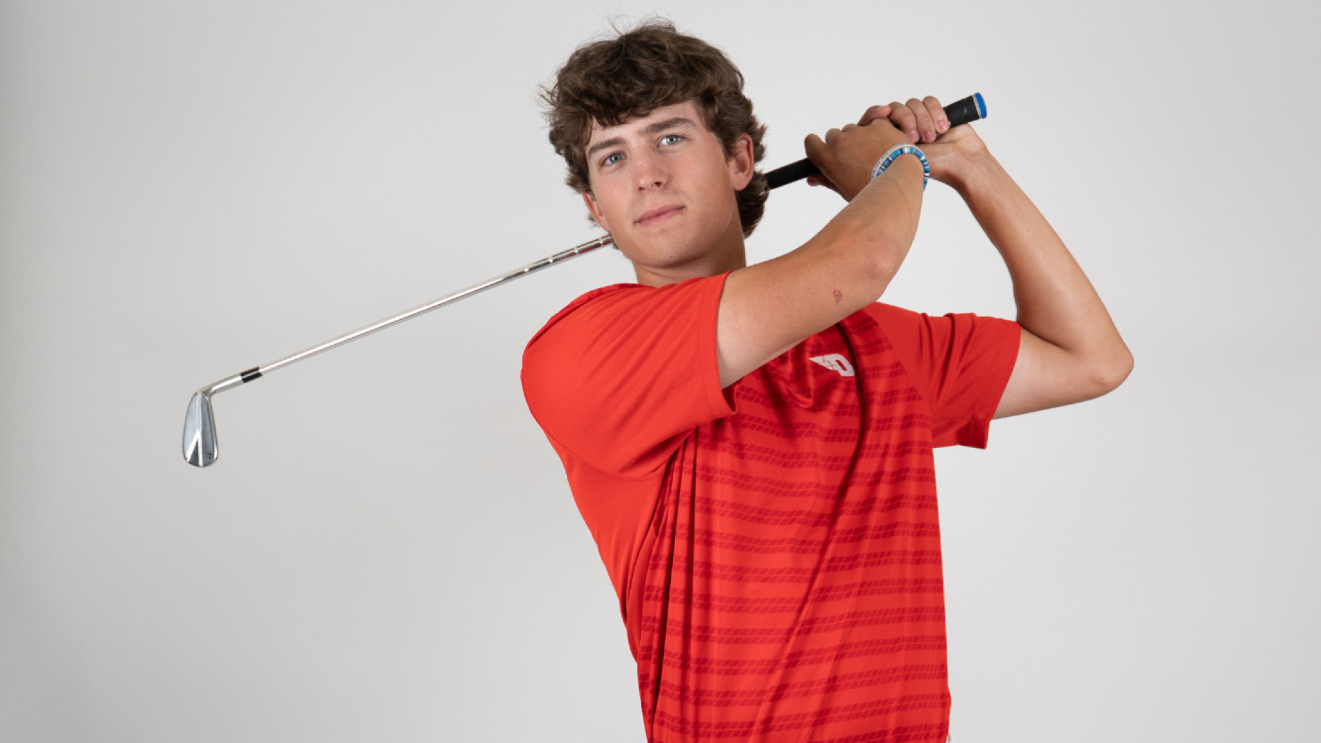 Men's golfer Victor Caliguri in his backswing with a club over his shoulder, wearing a red polo-style shirt with a white D logo, standing on a white background