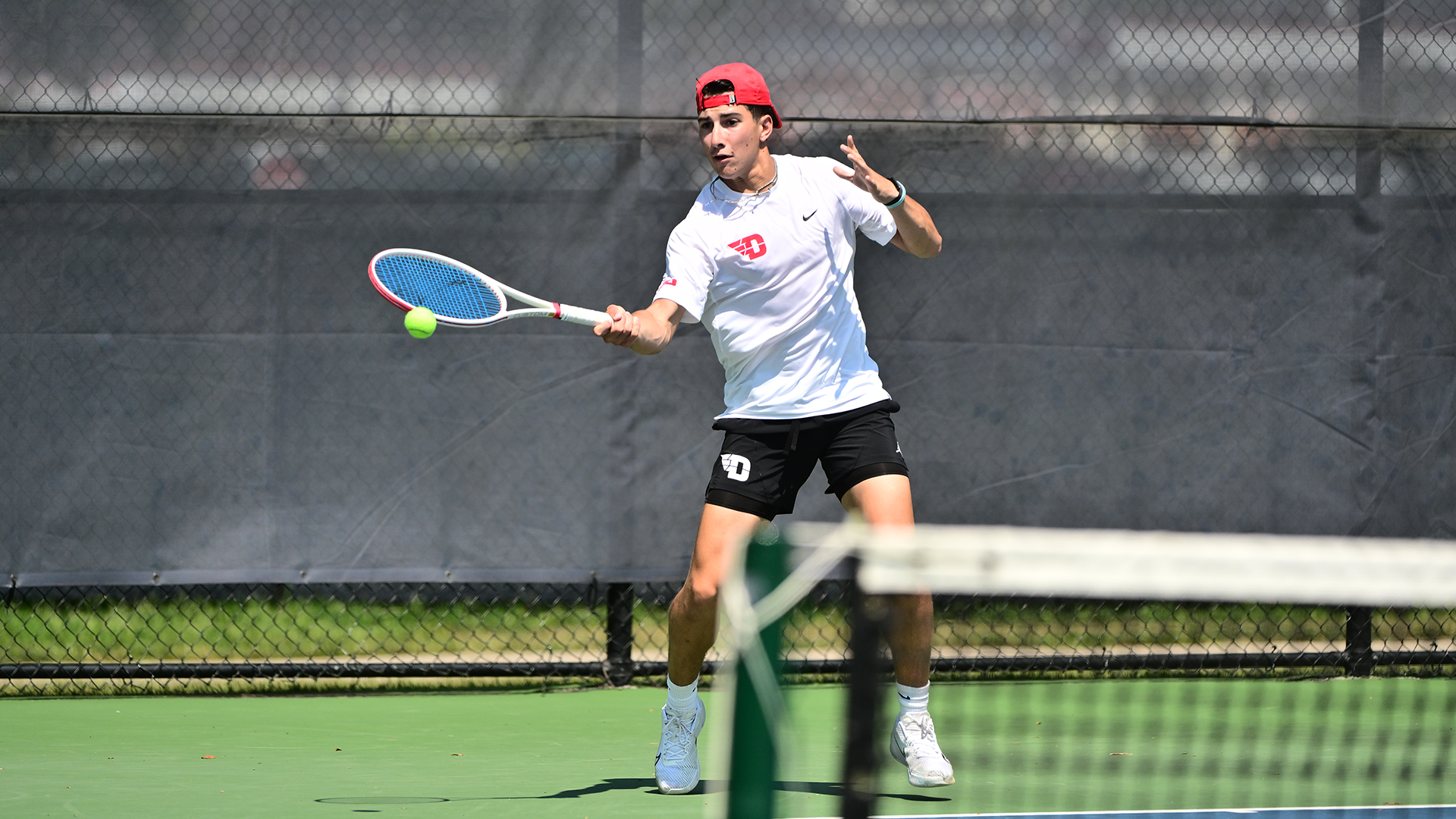 Kyle McNally slices a tennis ball, wearing a white shirt and black shorts.