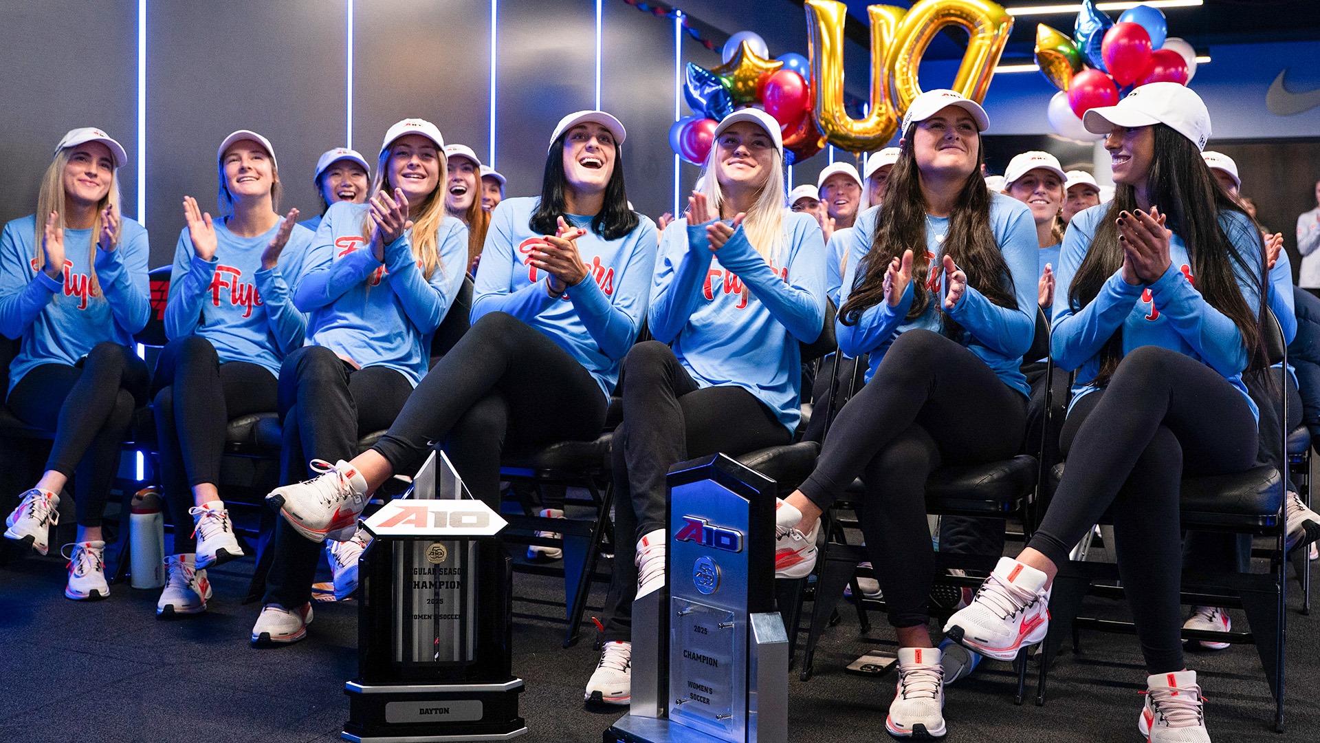 The Dayton women's soccer team claps while sitting behind the Atlantic 10 regular season and championship trophies.