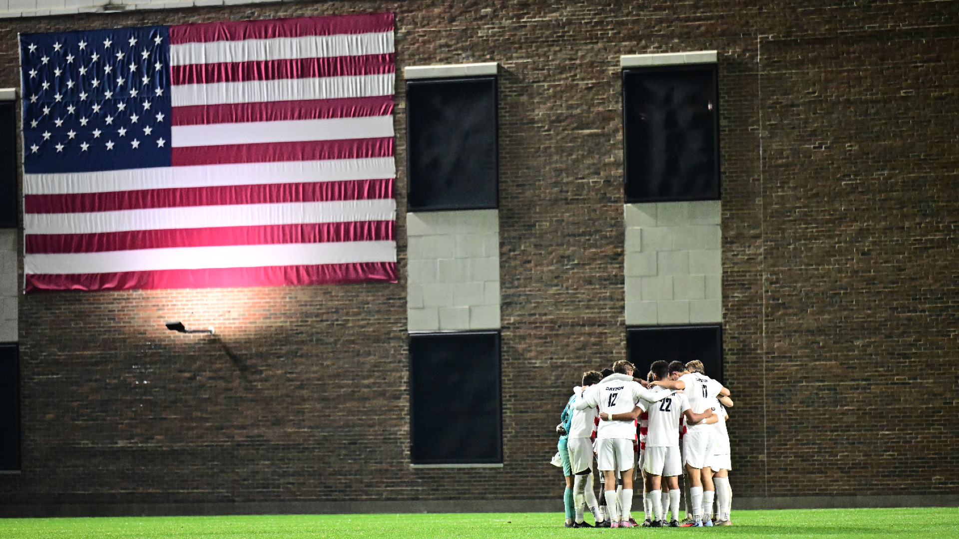 Members of the men's soccer team huddle on the field ahead of a match at Baujan Field. The American flag hangs on the building behind them and to the left.