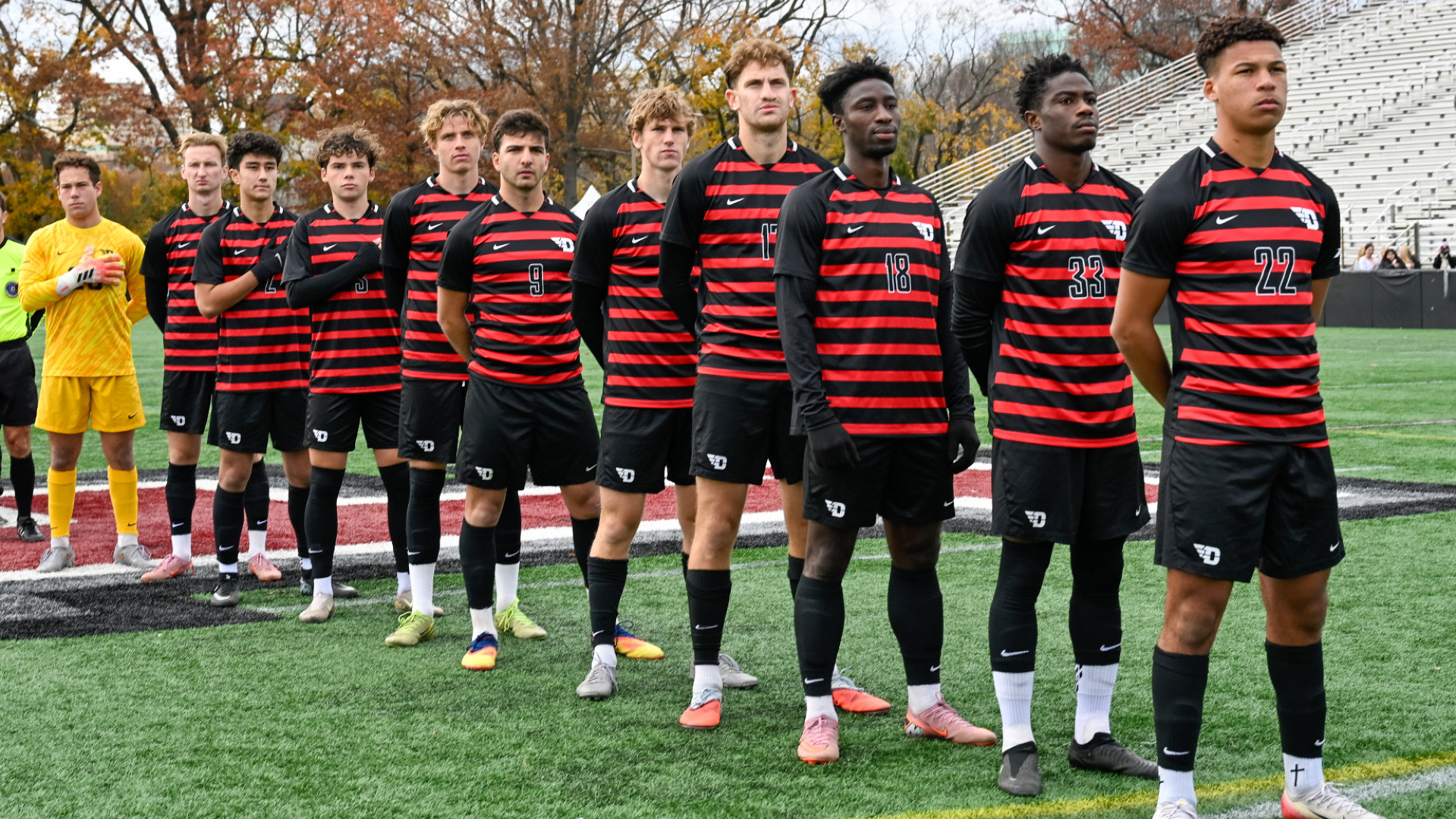 Members of the men's soccer team are lined up at centerfield, wearing red and black striped jersey tops and black shorts, during the National Athem ahead of their A-10 Semifinal match at Fordham