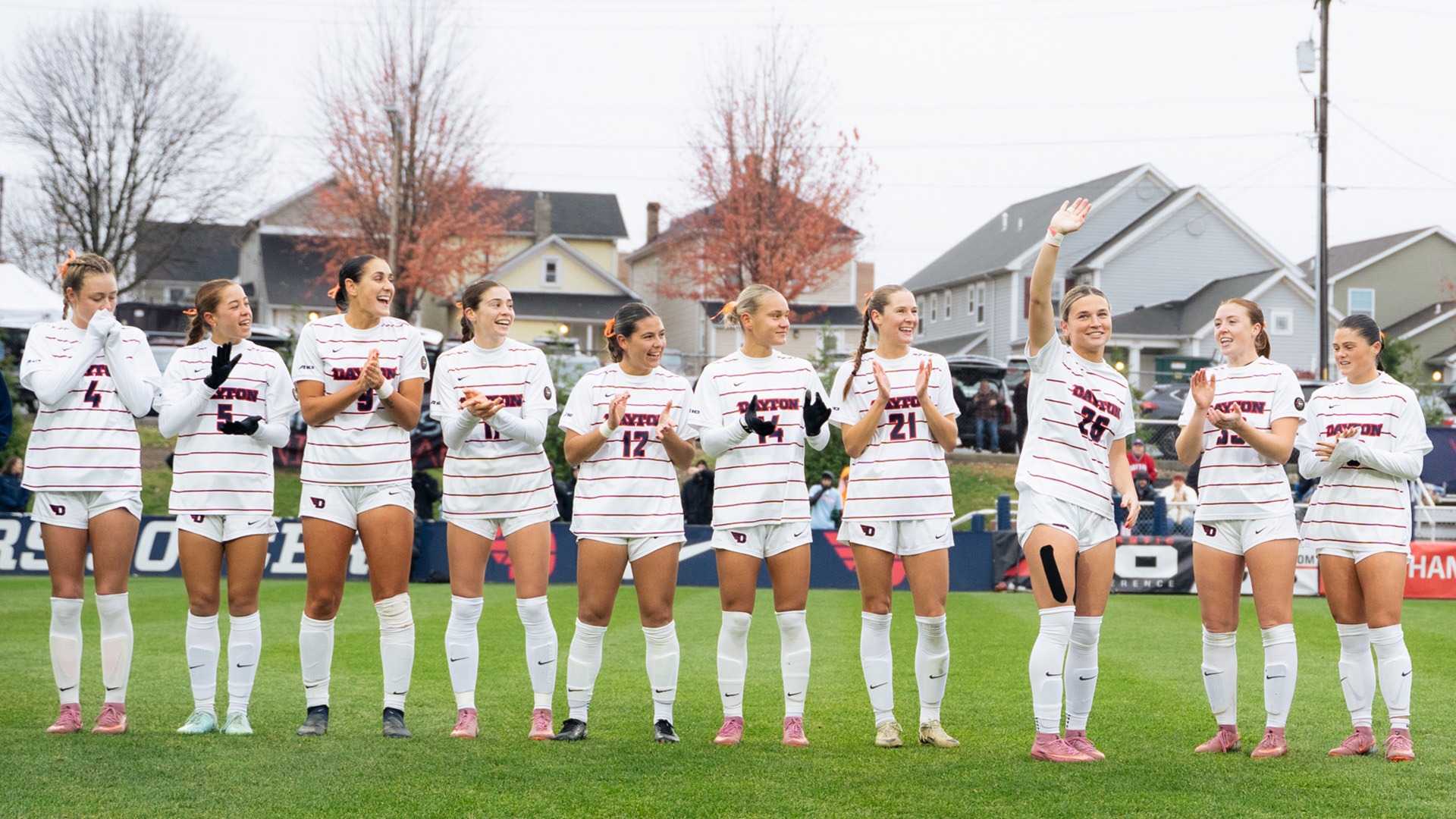 A row of Dayton women’s soccer players stands on the field during a pregame recognition, all wearing white uniforms with horizontal red stripes. The group is lined up shoulder-to-shoulder, smiling, clapping, and interacting with one another. One player near the right side raises her hand toward the crowd. The background shows fall trees, houses, and spectators gathered along the sideline on an overcast day.