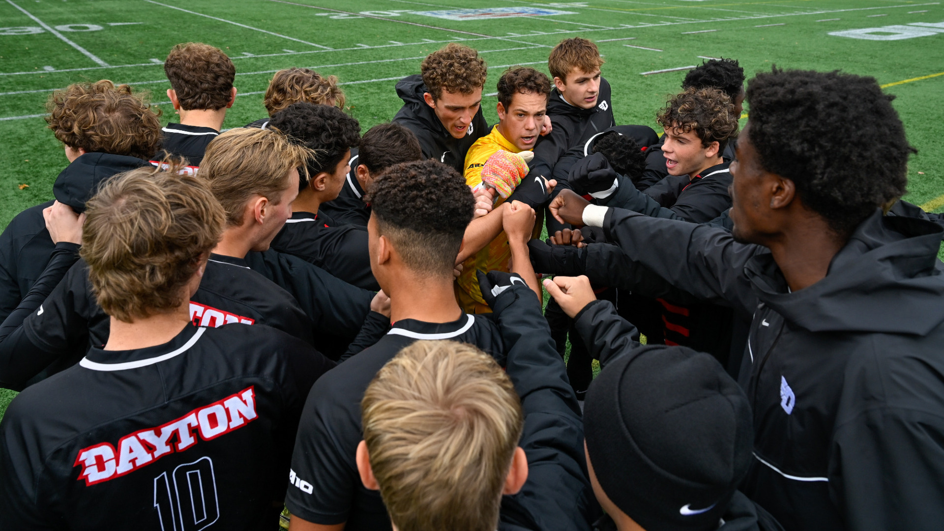 Members of the men's soccer team huddle together, all wearing black except the goalkeeper who has on a yellow jersey, with one hand in the center ahead of the team's A-10 semifinal match at Fordham