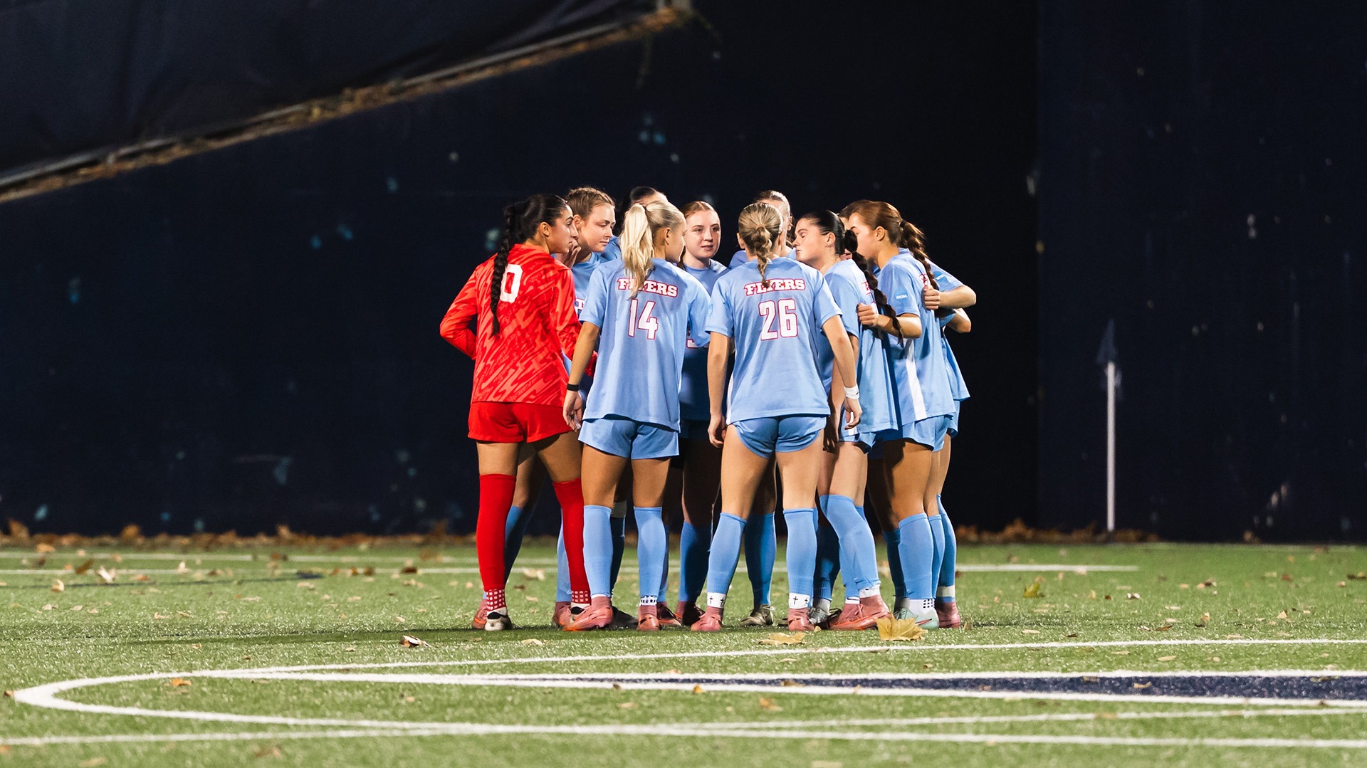 A huddle of Dayton women’s soccer players stands together on the field before kickoff at night. The team, wearing light blue uniforms with “Flyers” on the back, forms a tight circle with arms around each other. The goalkeeper in a red kit joins the group. Fallen leaves are scattered across the turf as stadium lights illuminate the scene against a dark background.