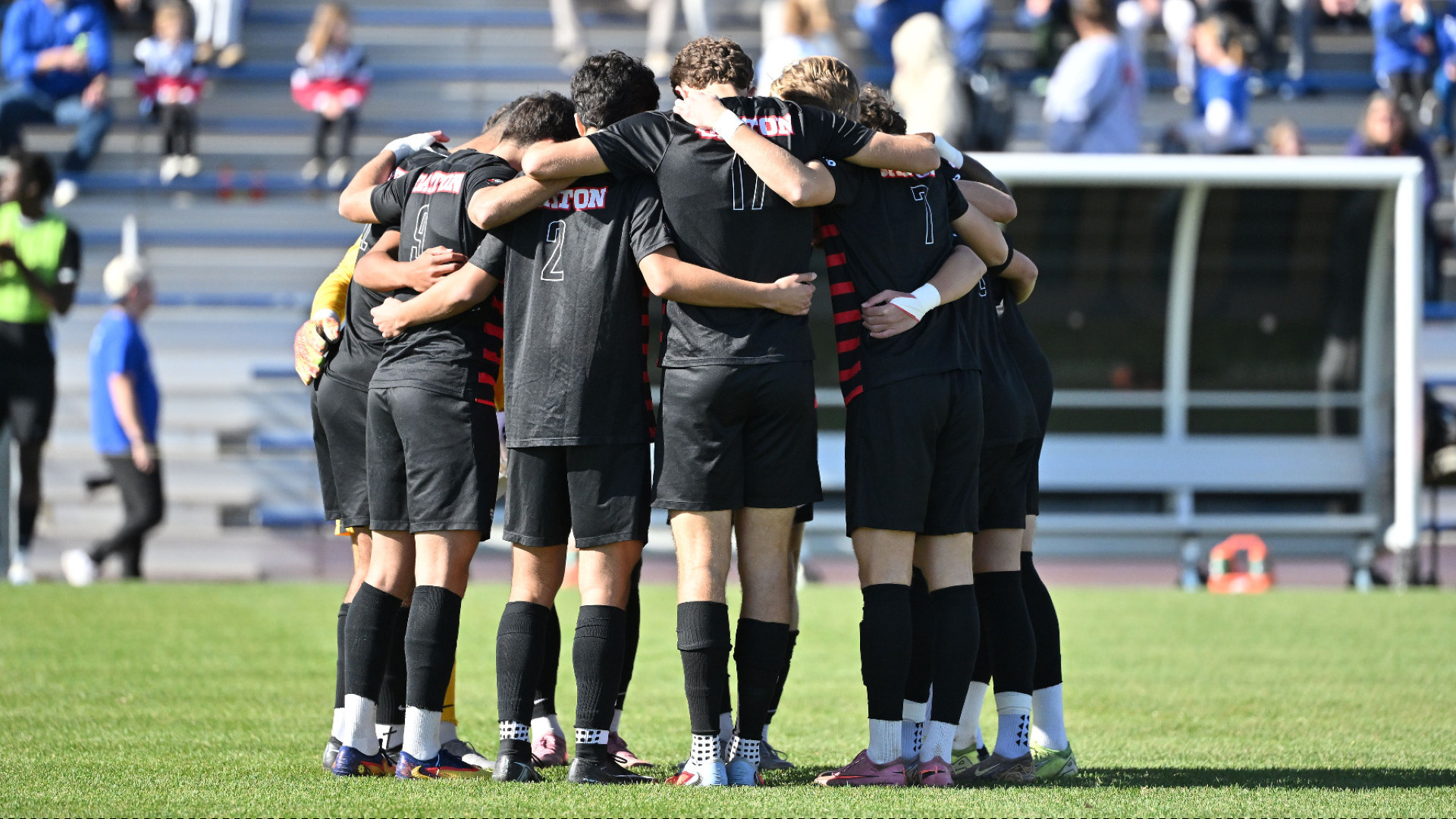 Members of the men's soccer team, wearing black jersey tops and black shorts, huddle together on the field before the team's Atlantic 10 Championship final match at Saint Louis