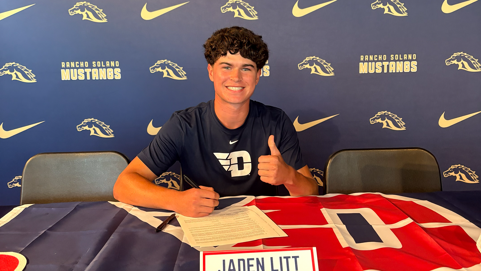 Jaden Litt sits at a table that has a Dayton Flyers flag and other Dayton decorations on it. He's giving a thumbs up with his left hand and smiling.