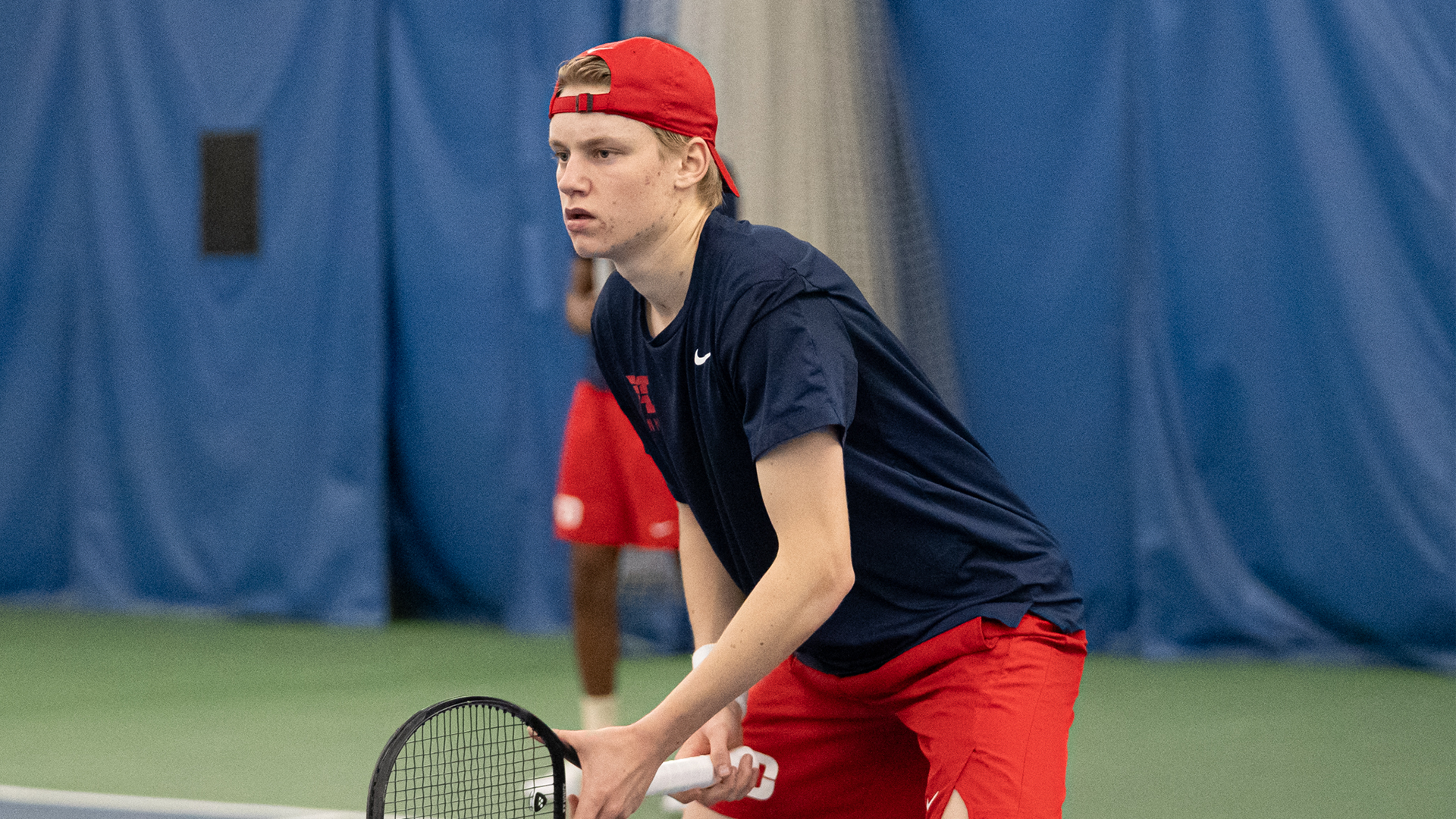 Andreas Nygaard stands hunched and ready to play, holding his racquet in his right hand and supporting it with his left.