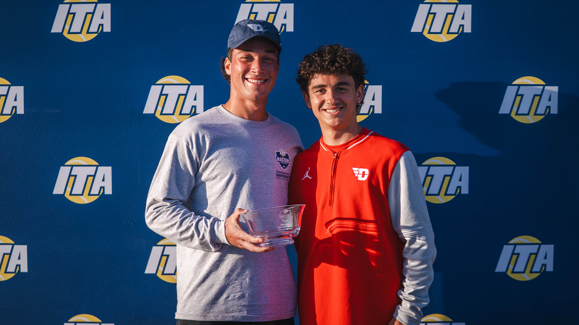 Mikkel Zinder and Dylan Jaen pose for a picture in front of an ITA Conference Masters blue backdrop. Zinder is holding his trophy for being the back draw co-champion.