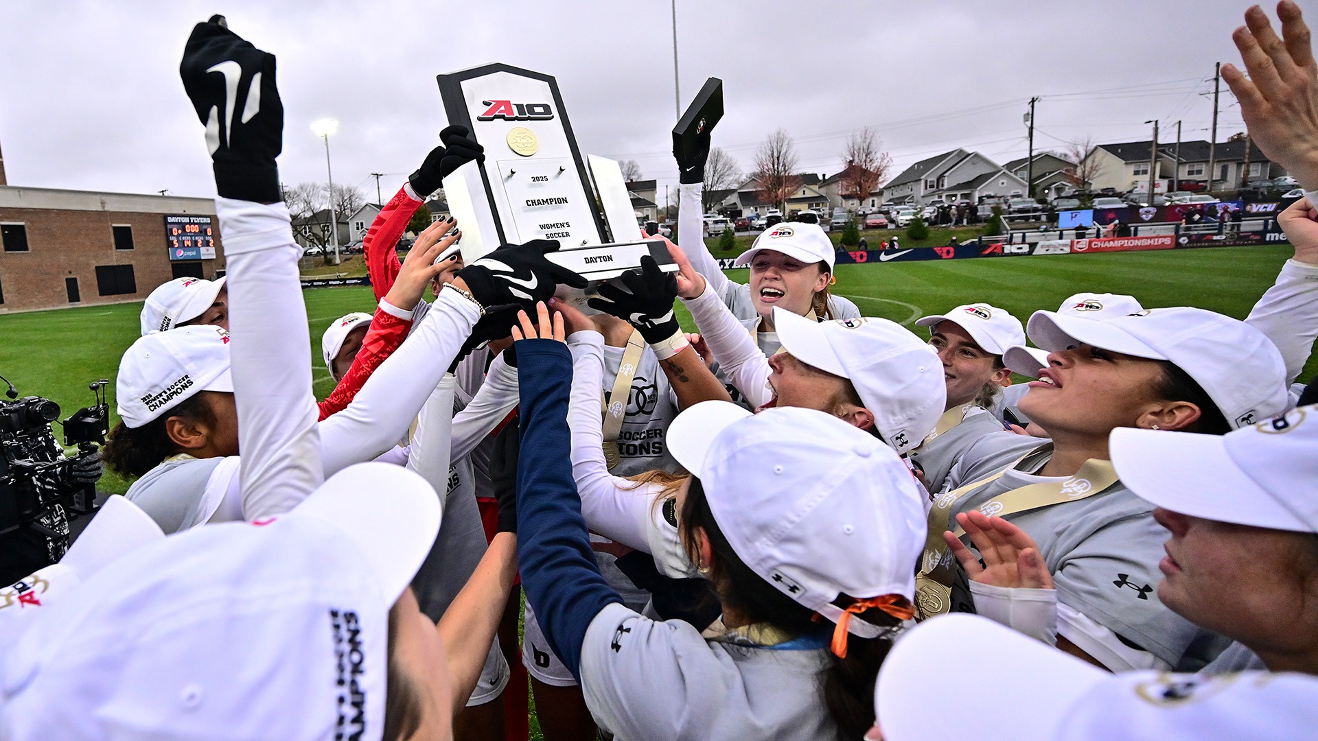 Dayton women’s soccer players celebrate together on the field, raising the 2025 Atlantic 10 Championship trophy high above their heads. The team, wearing white hats and gloves, smiles and cheers in a joyful huddle under cloudy skies after clinching the conference title.