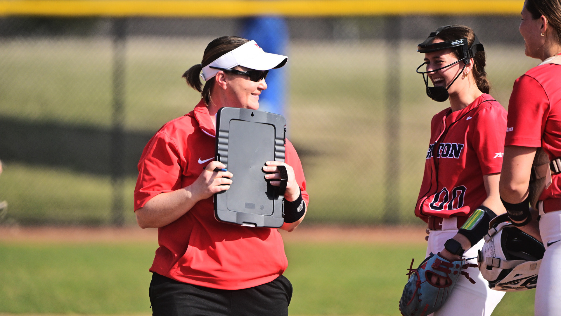 Softball head coach Cara Clark, wearing a red top and white visor hat and holding a black clipboard, stands on the field talking with pitcher and catcher during a game at UD Softball Stadium against Stonehill 3-12-25 ES