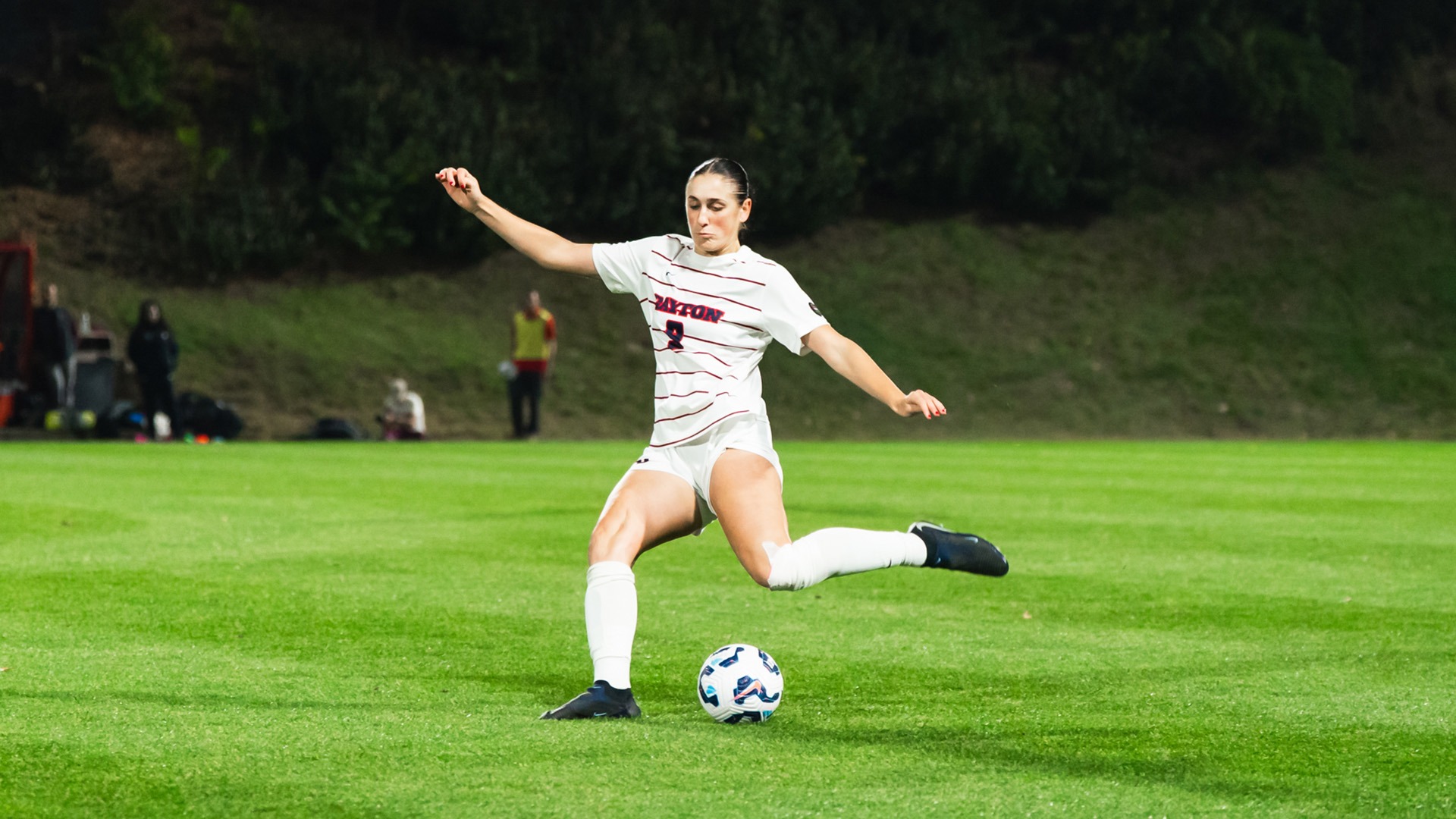 University of Dayton women’s soccer player Ella Raimondi winds up and strikes the ball with her right foot on a green grass field during a match, wearing a white Flyers uniform with red accents and the number 9, with teammates and the bench visible in the background at night.