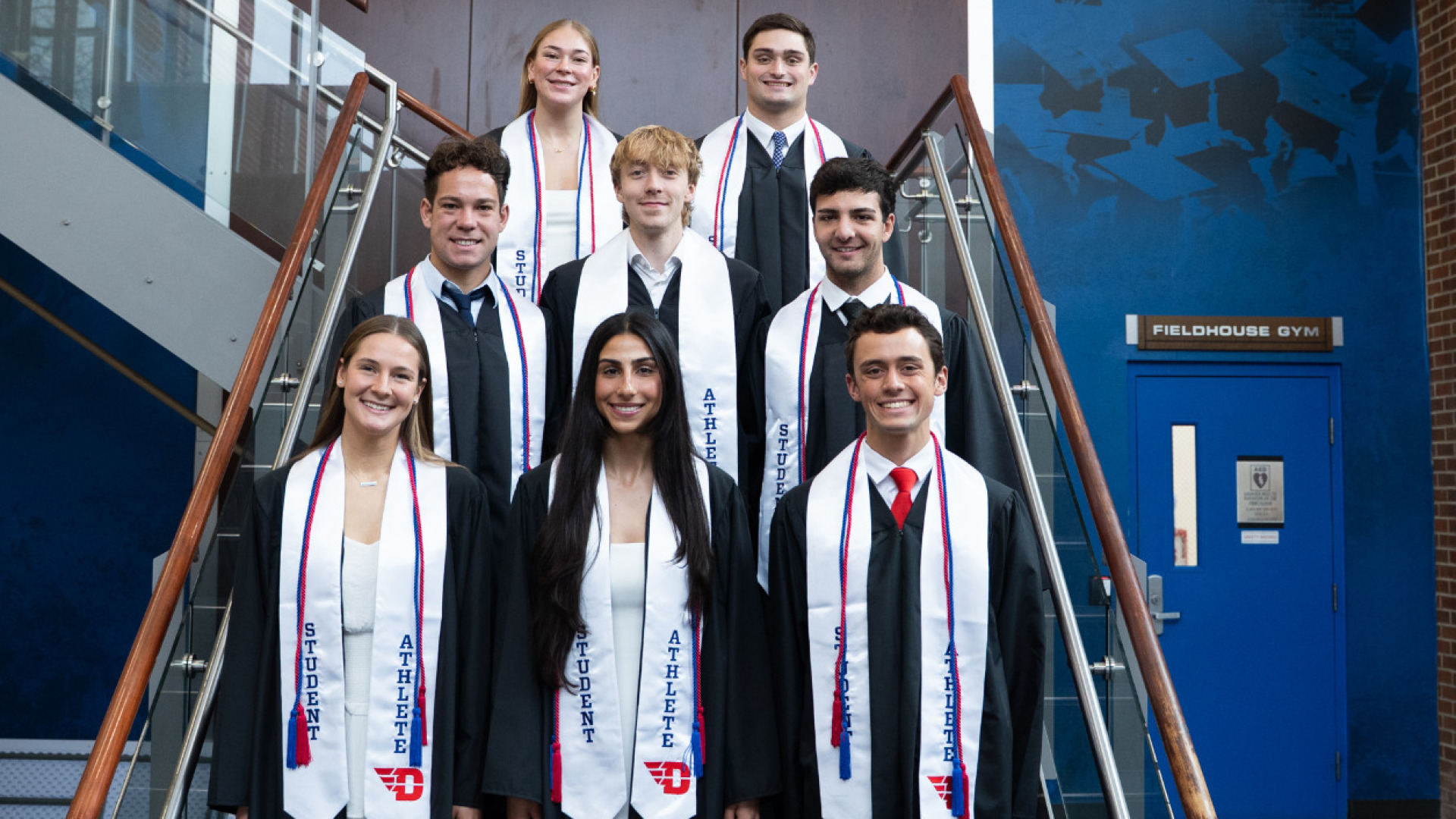 Several student-atheletes dressed in graduation gowns posed together on the stairwell in Cronin Athletic Center ahead of December 2025 graduation