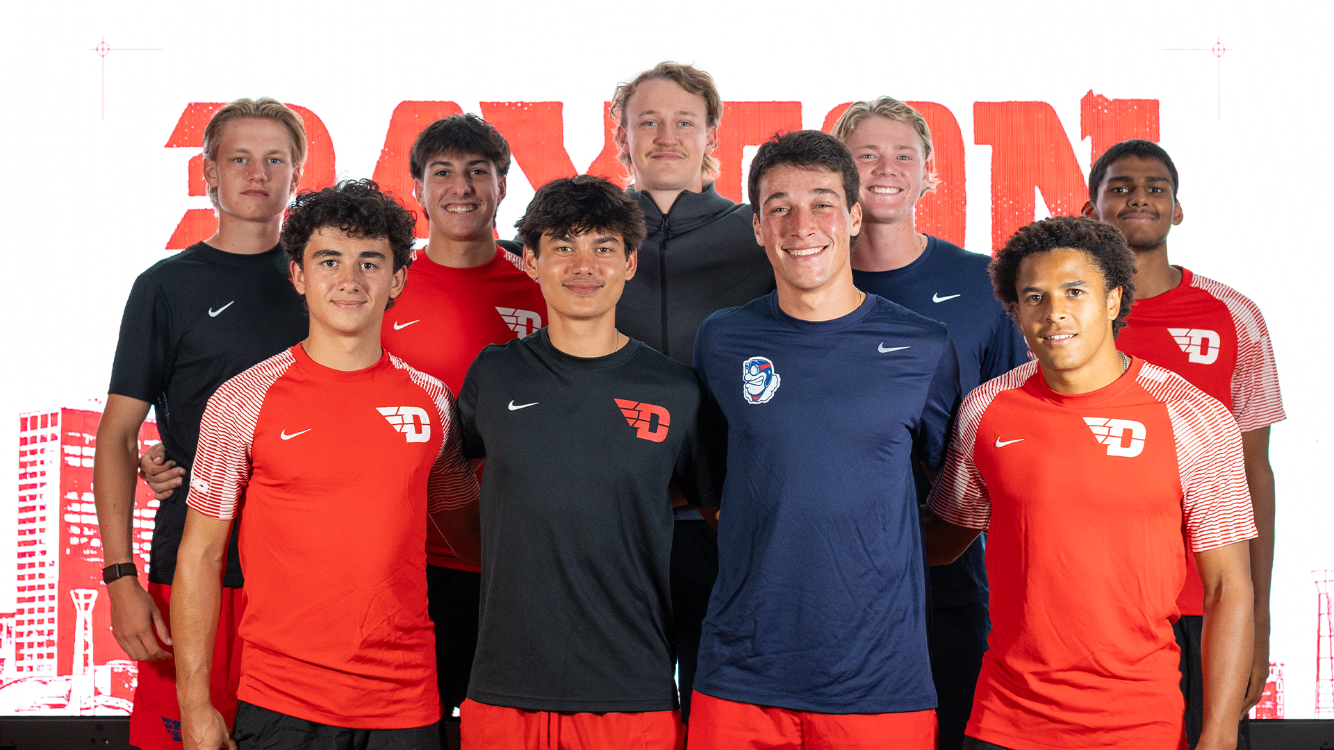 The men's tennis team poses for a team picture in front of a white and red Dayton background. The players are all in different Dayton gear.