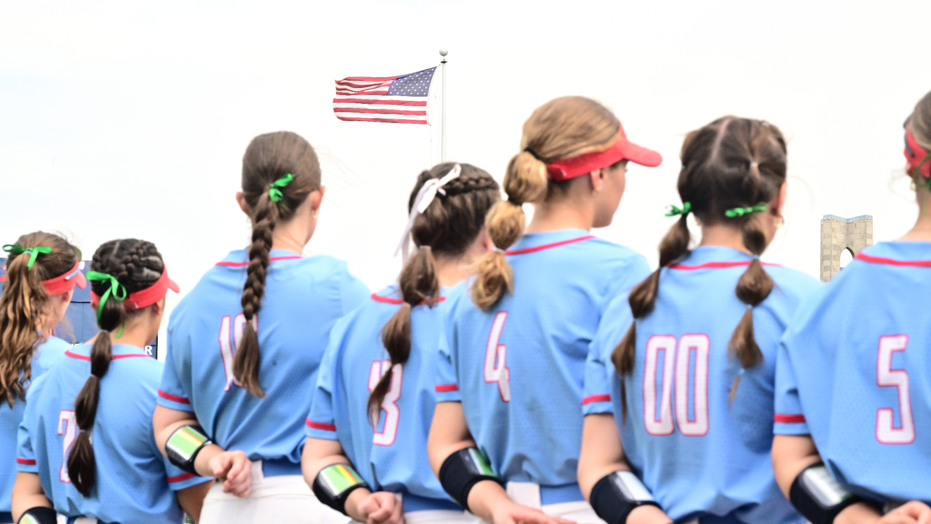Members of the softball team standing for the national athem with the American flag flying in the distance at UD Softball Stadium