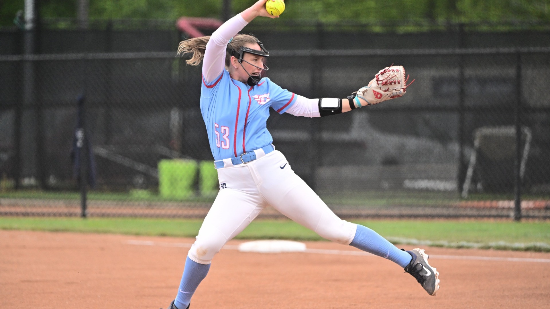 Softball right-handed pitcher Izzy Kemp shown from the side with the ball at the top of her throwing motion wearing a Chapel blue uniform top and white pants